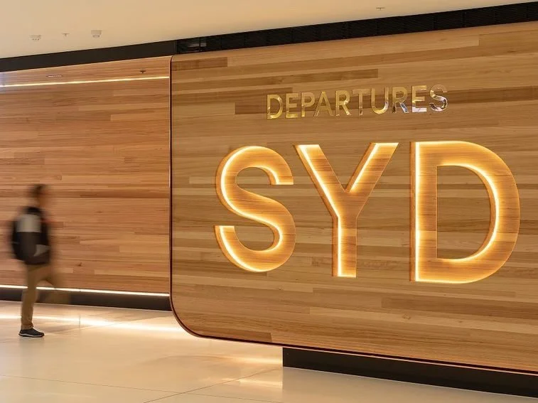 Inside an airport terminal, a large wooden wall with illuminated gold letters spelling "SYD" and smaller gold letters spelling "DEPARTURES" above it. A person with a backpack walks past.