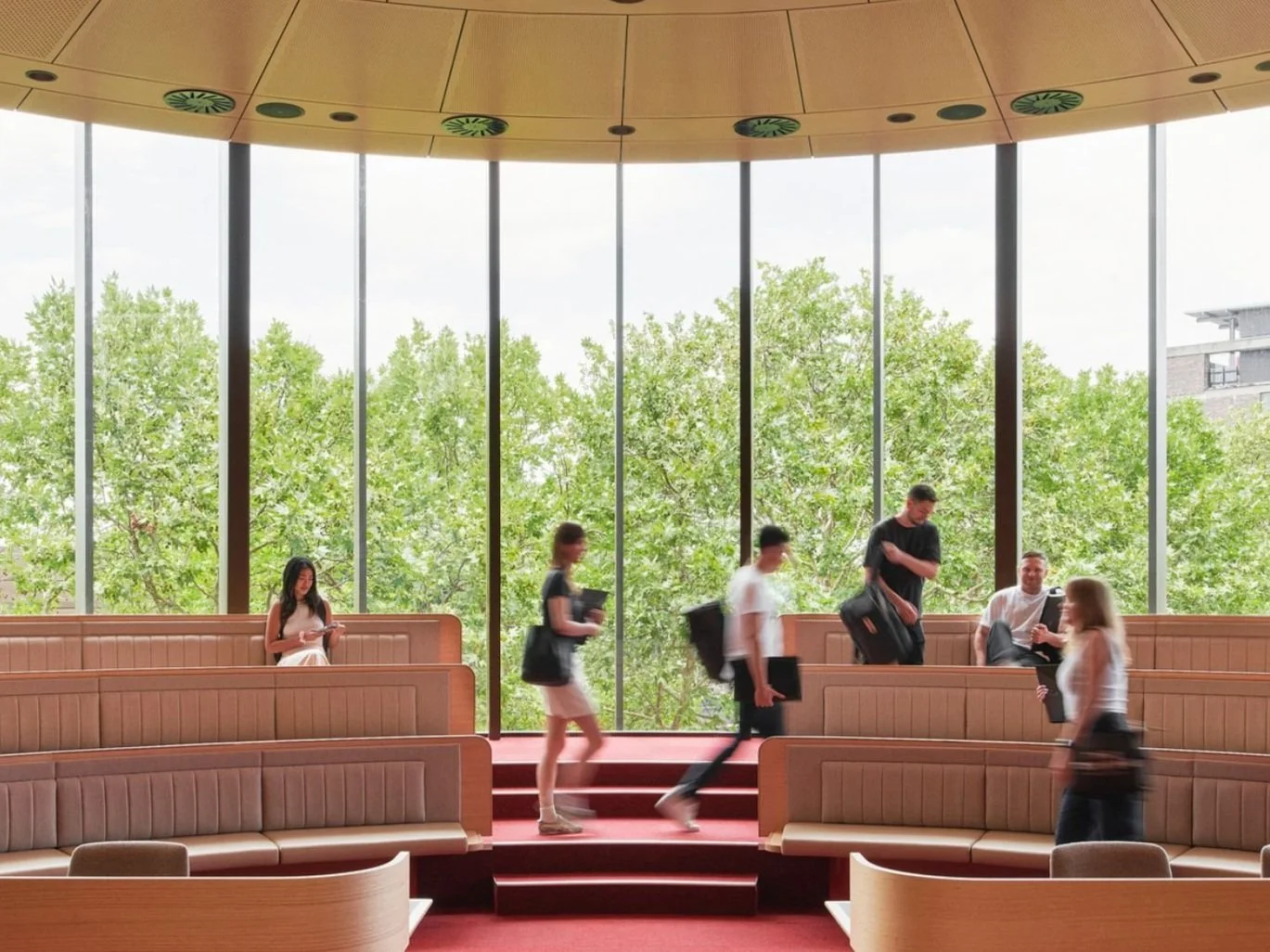 People sitting and walking inside a modern waiting area with large windows and green trees outside.