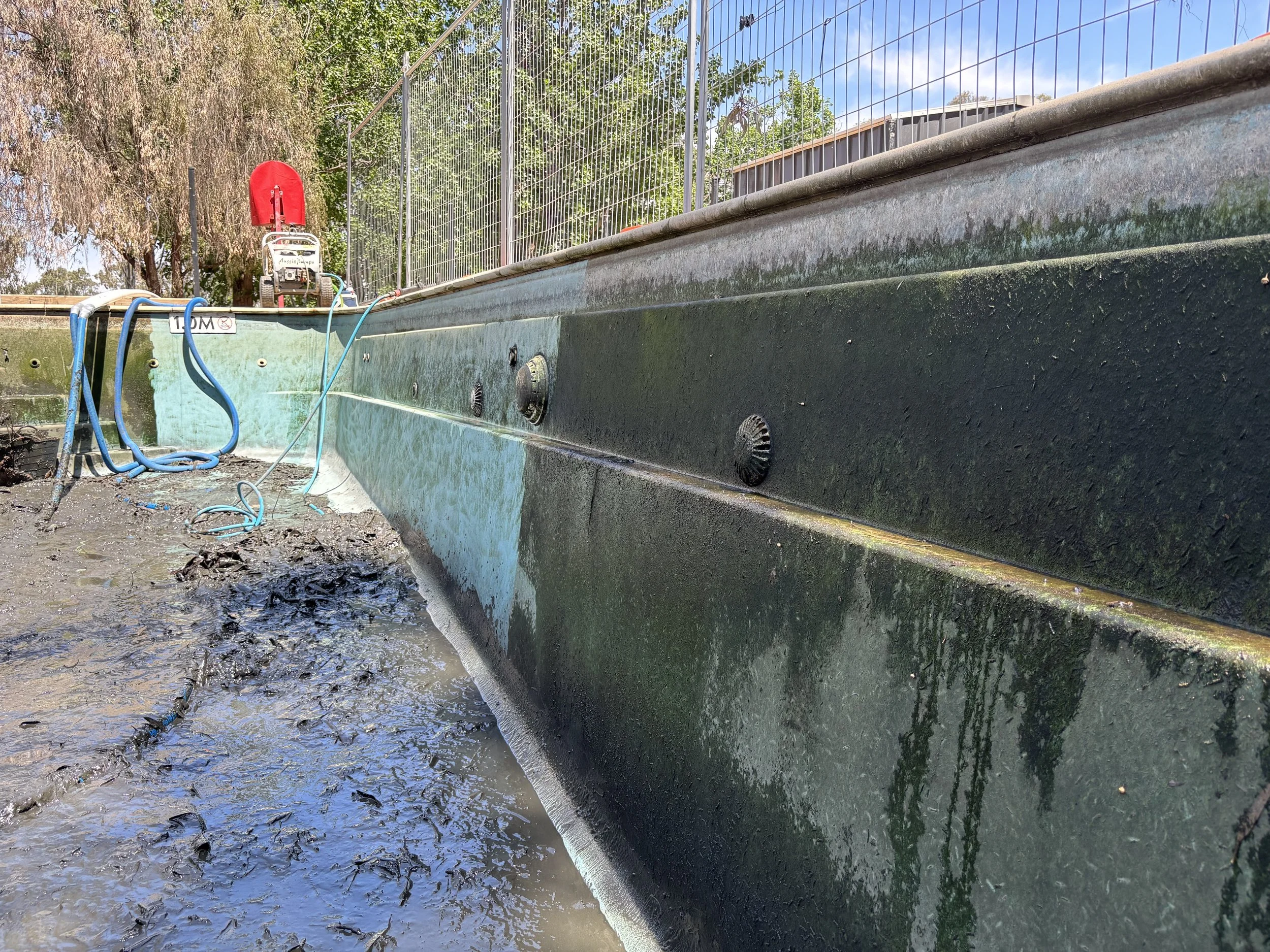 The image shows the side of a boat being repaired or cleaned, with water, mud, and tools around it. There are hoses and a red sprayer on top, and the boat is in a water body. The background has trees and a blue sky.