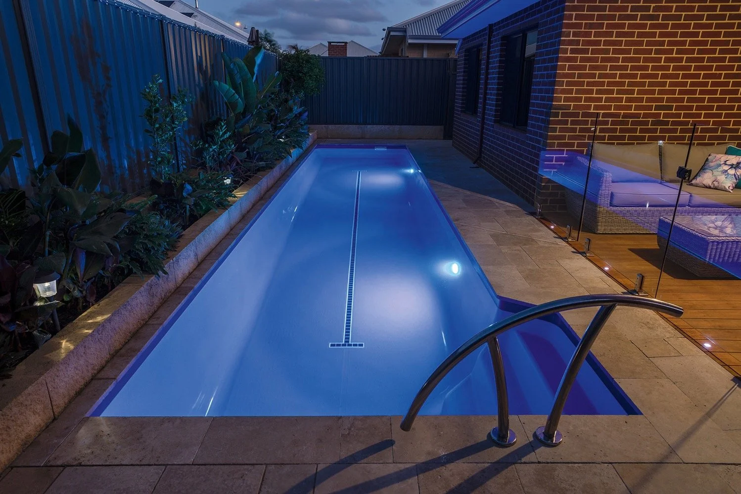 A backyard with a narrow illuminated swimming pool at night, bordered by plants and a brick wall, with a glass fence on one side and a patio with outdoor furniture.