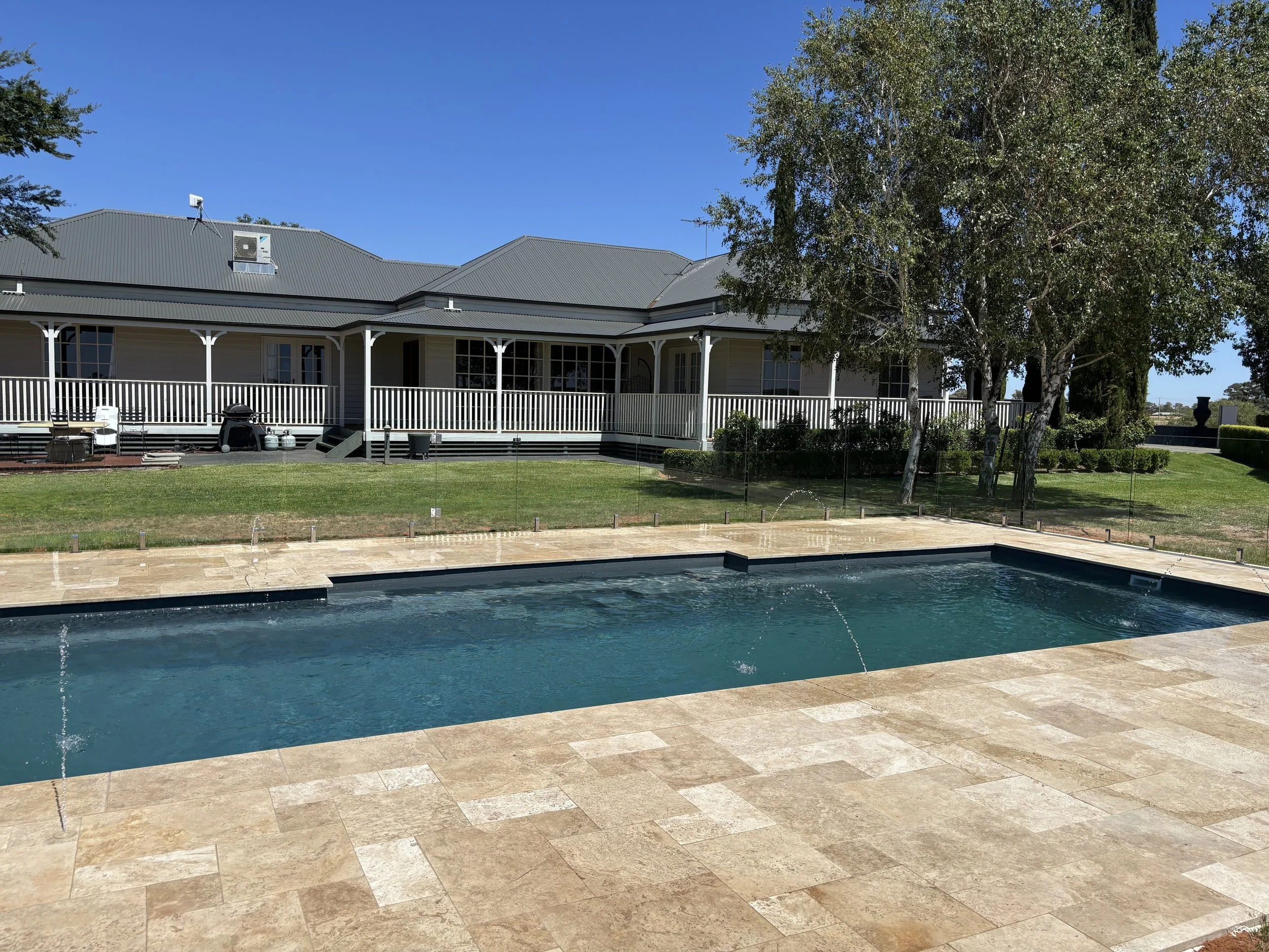 A backyard featuring a swimming pool with water jets, a stone deck, a grassy lawn, and a large house with a wrap-around porch and trees.