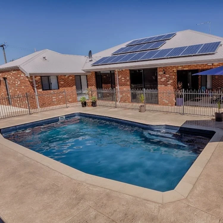 Backyard with a small in-ground swimming pool surrounded by a concrete deck, fenced patio area, brick house with solar panels on the roof, clear blue sky.