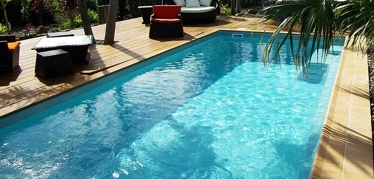A backyard swimming pool surrounded by a wooden deck with outdoor seating and tropical plants.