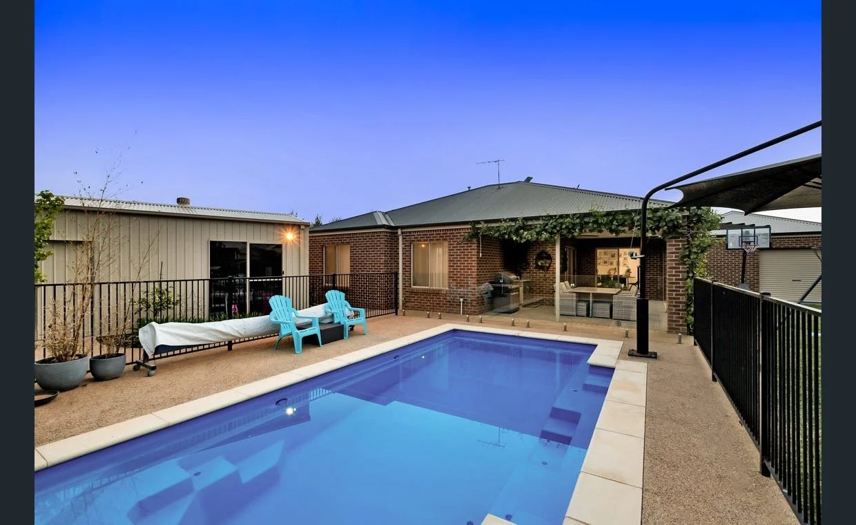 Backyard with a swimming pool, two blue chairs, potted plants, a covered patio with outdoor furniture, and a basketball hoop during twilight.
