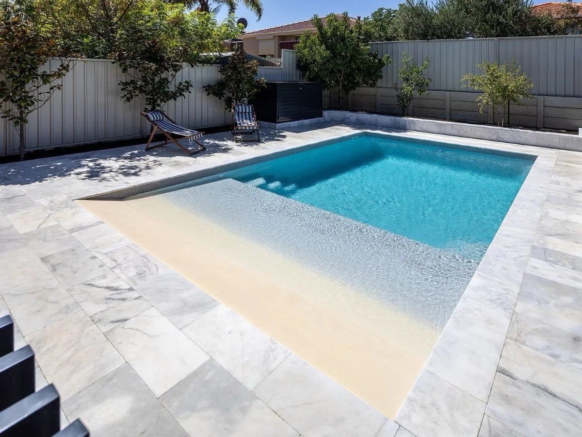 A backyard swimming pool with a white and beige textured pool edge, surrounded by a marble-tile patio, with lounge chairs and trees along a gray fence.