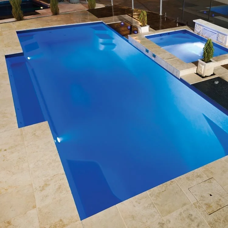 An outdoor swimming pool with bright blue water, illuminated by underwater lights, surrounded by a stone deck. Two smaller hot tubs are visible in the background, each with plants in planters on their edges. A glass railing encloses the area.