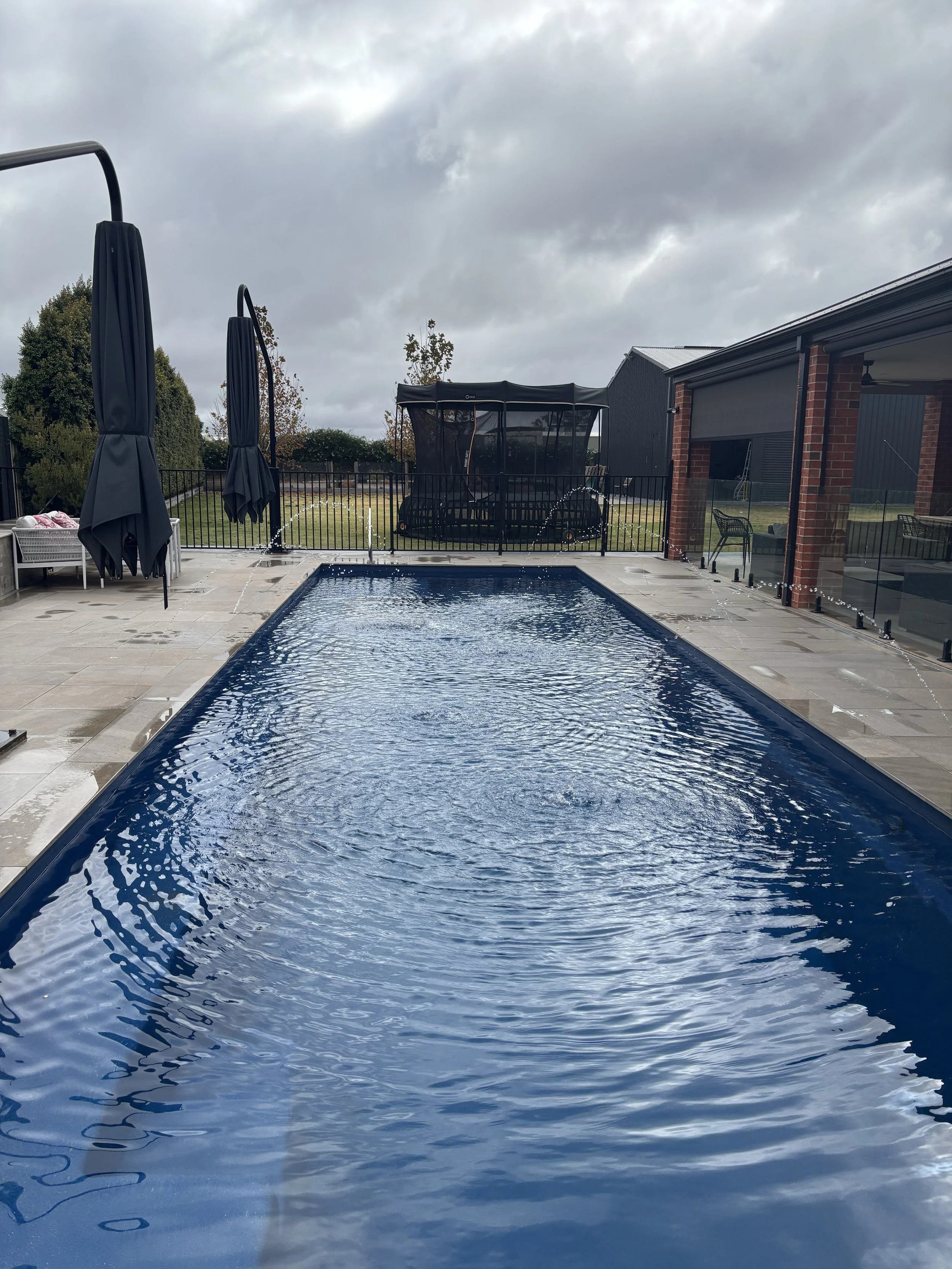 Backyard scene with a rectangular swimming pool, two black umbrellas, patio furniture, a trampoline, and a cloudy sky.