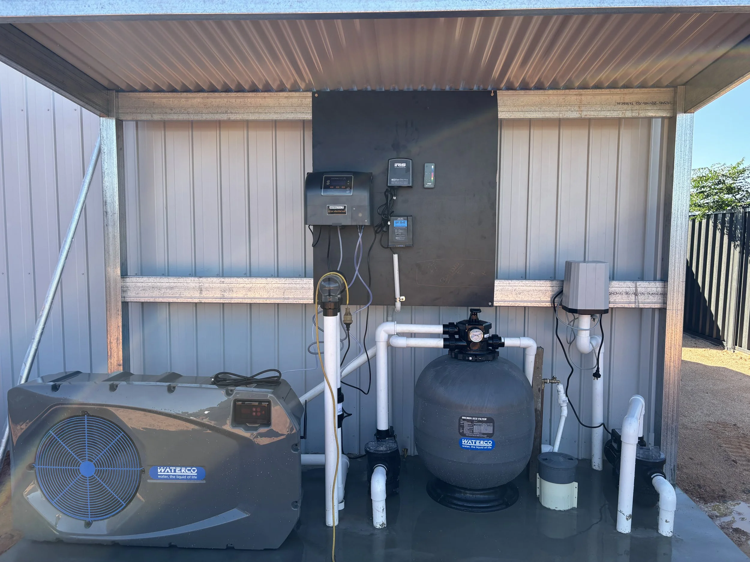A water treatment system with a water filter tank, a water pump, and control units mounted on a black panel in an outdoor shelter with a corrugated metal roof and walls.