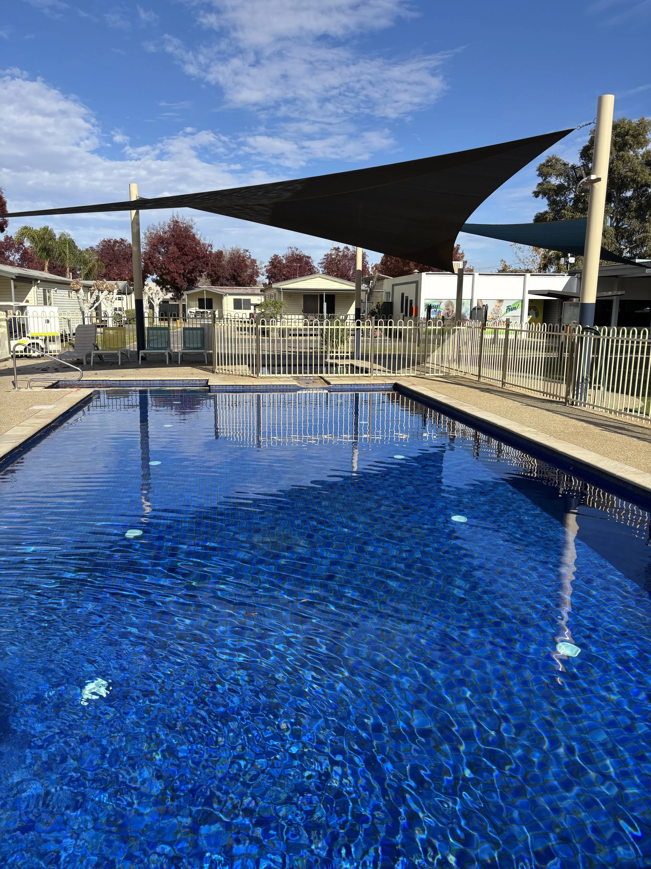 Swimming pool with blue tiles, surrounded by a fence, with pool chairs and shaded canopies in a community or residential area, under a partly cloudy sky with some trees and houses in the background.
