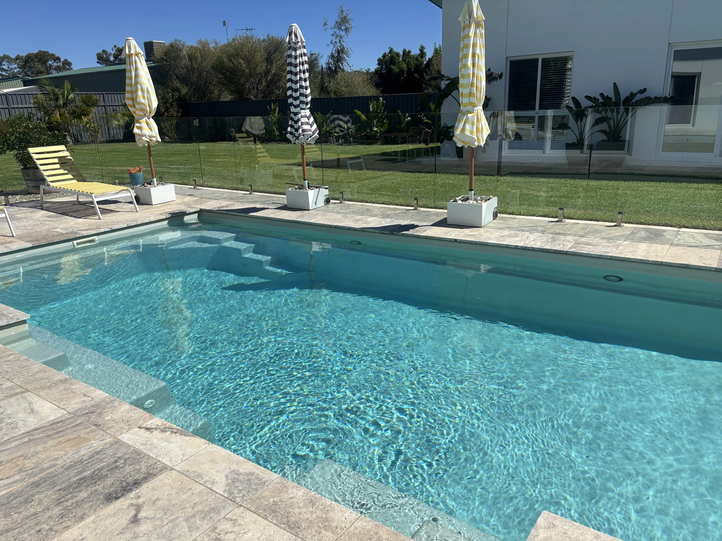 Clear swimming pool with steps at the shallow end, surrounded by a tiled deck, with yellow and white striped umbrellas and lounge chairs on a sunny day. There is a glass fence around the yard, grass, trees, and a modern house in the background.