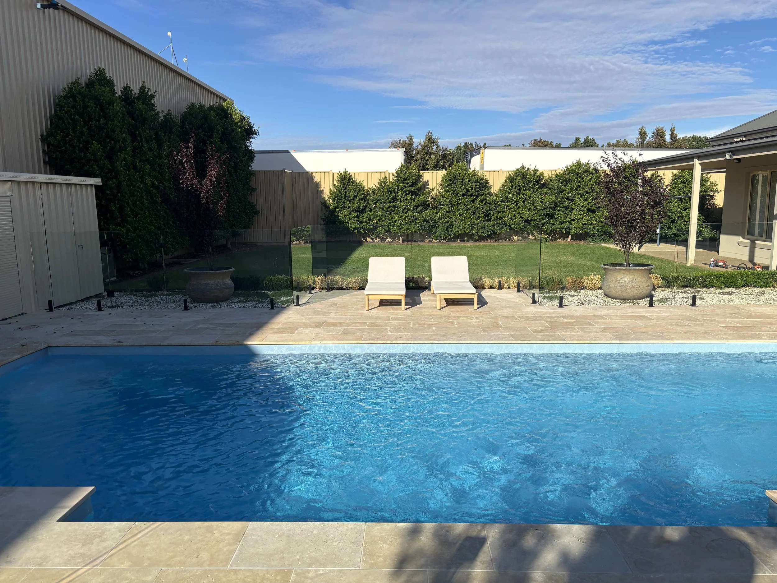 A backyard swimming pool with two white lounge chairs on a stone patio, green bushes, and trees with a blue sky overhead.