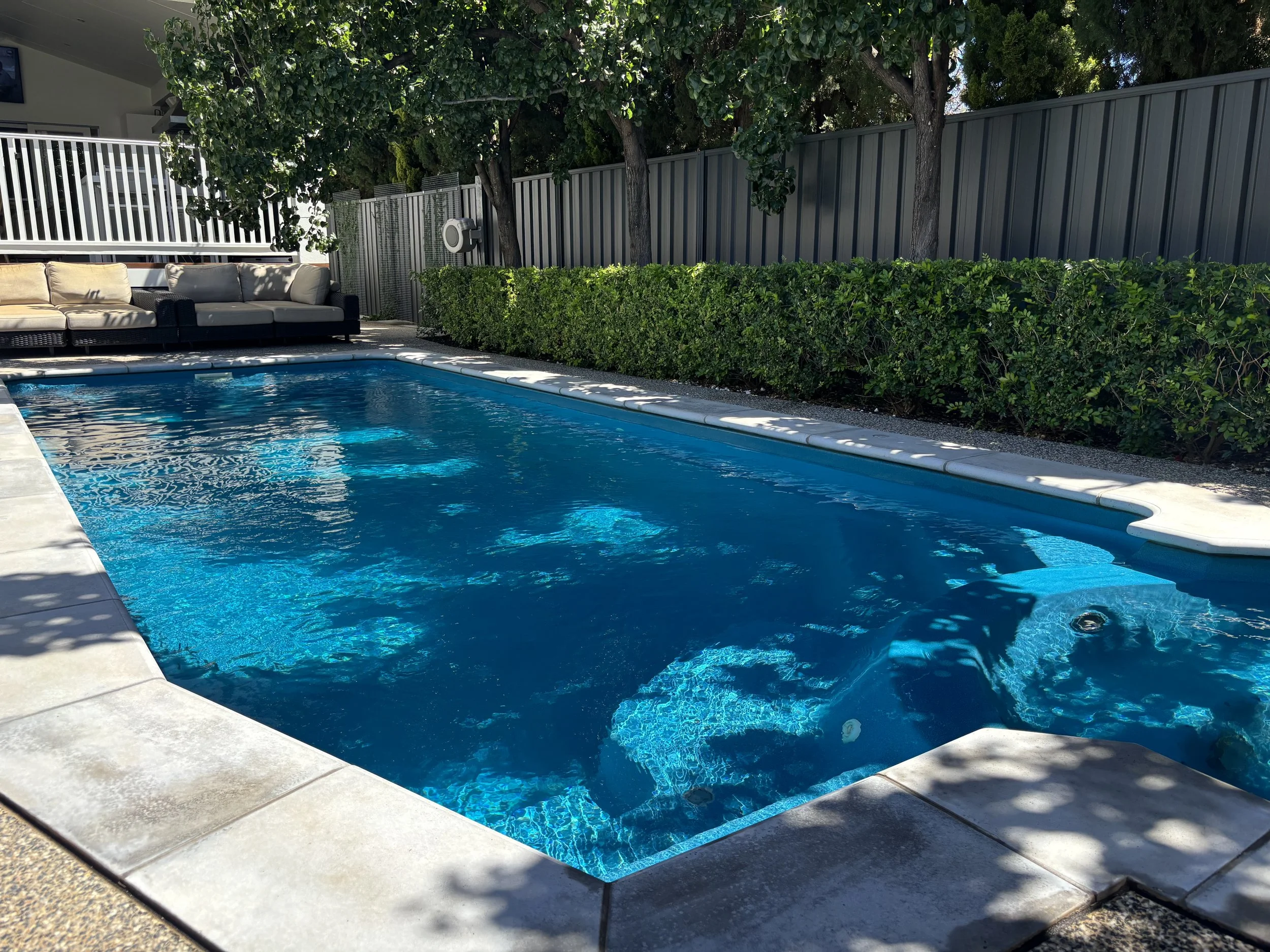 A backyard swimming pool with clear blue water, surrounded by a concrete deck, with outdoor seating and green hedges along a gray metal fence, shaded by trees.