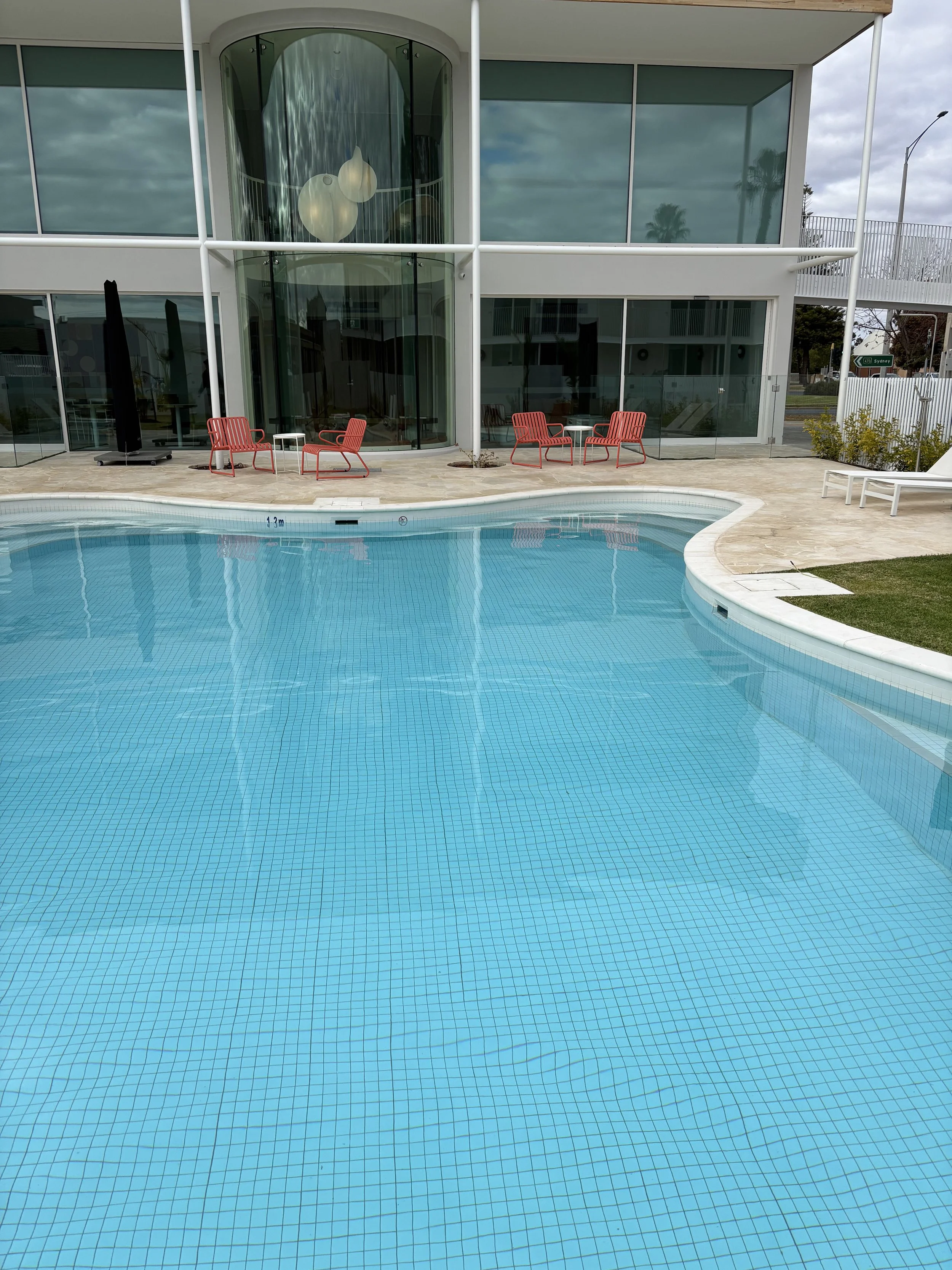 Empty swimming pool with blue tiles in front of a modern glass building, with red lounge chairs and small tables on the pool deck.