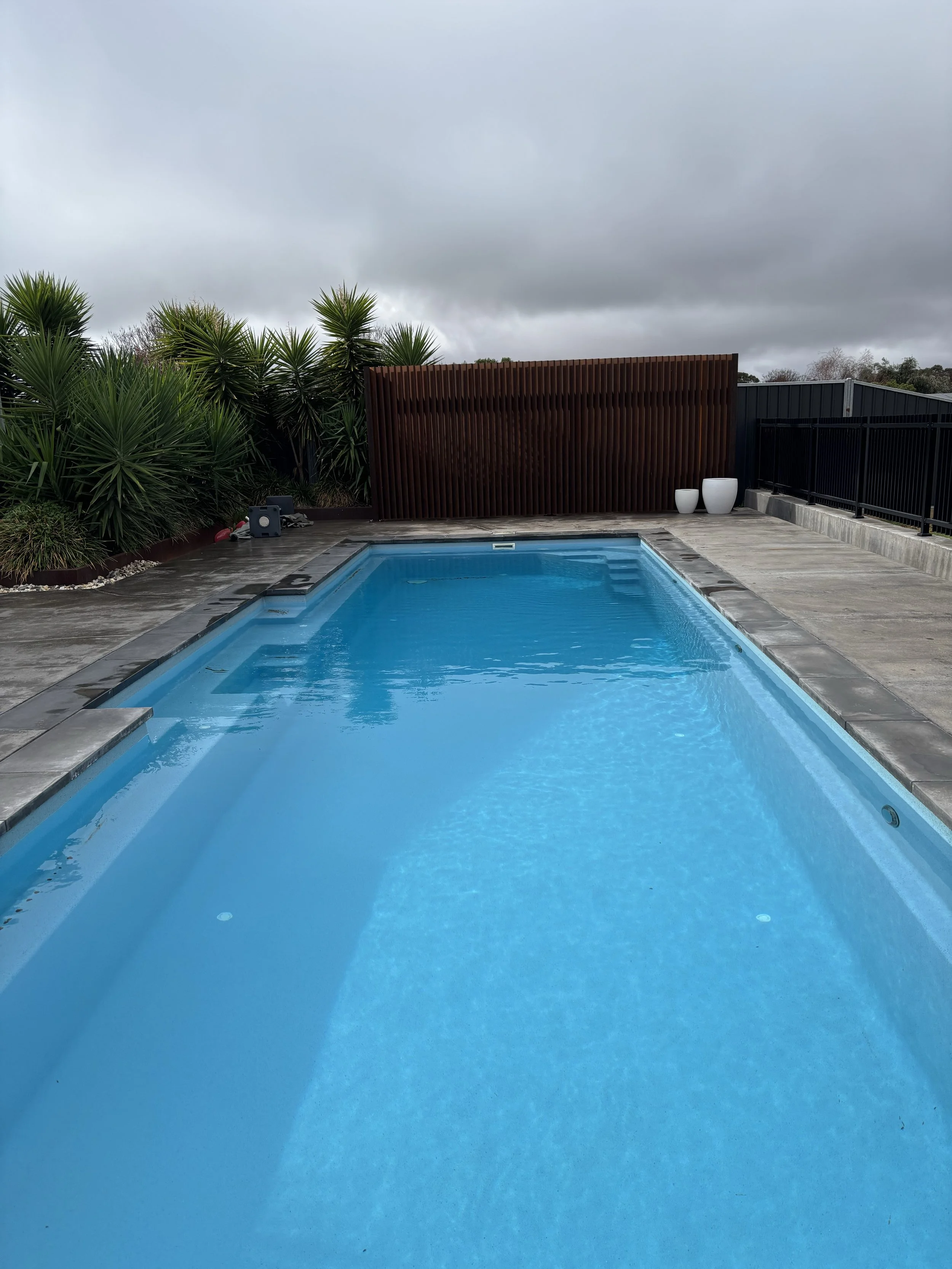 An empty rectangular swimming pool with blue water, surrounded by a concrete deck, with greenery and a brown wooden privacy screen in the background under a cloudy sky.
