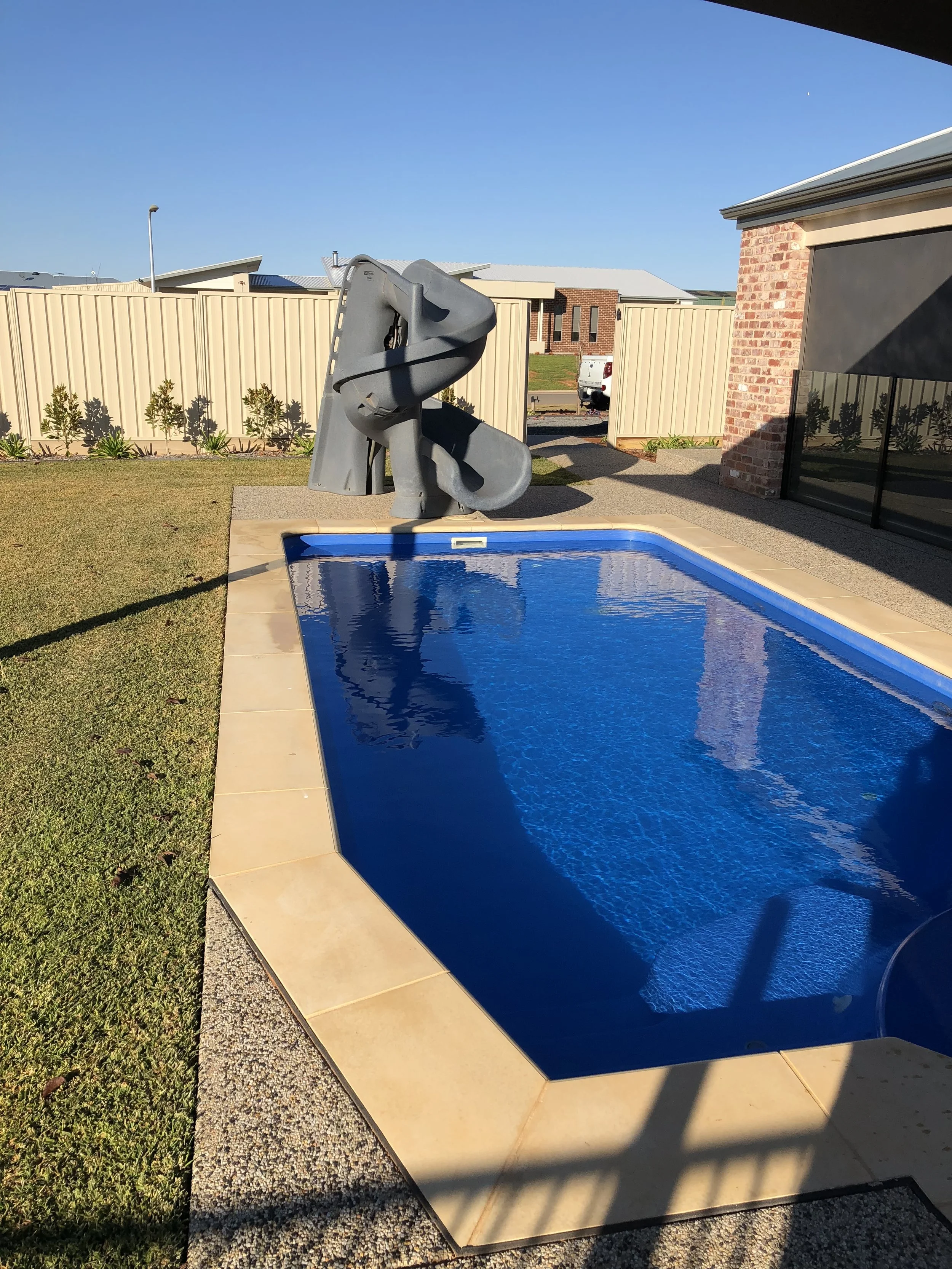 A backyard swimming pool with a water slide next to a house, surrounded by a lawn and fenced backyard under a clear blue sky.