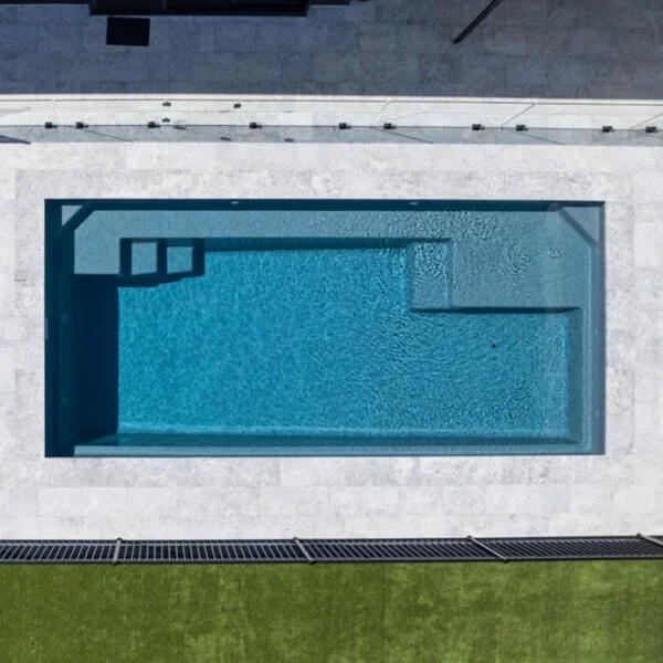 Aerial view of a rectangular swimming pool with blue water, surrounded by a light gray deck, and a green lawn at the bottom.