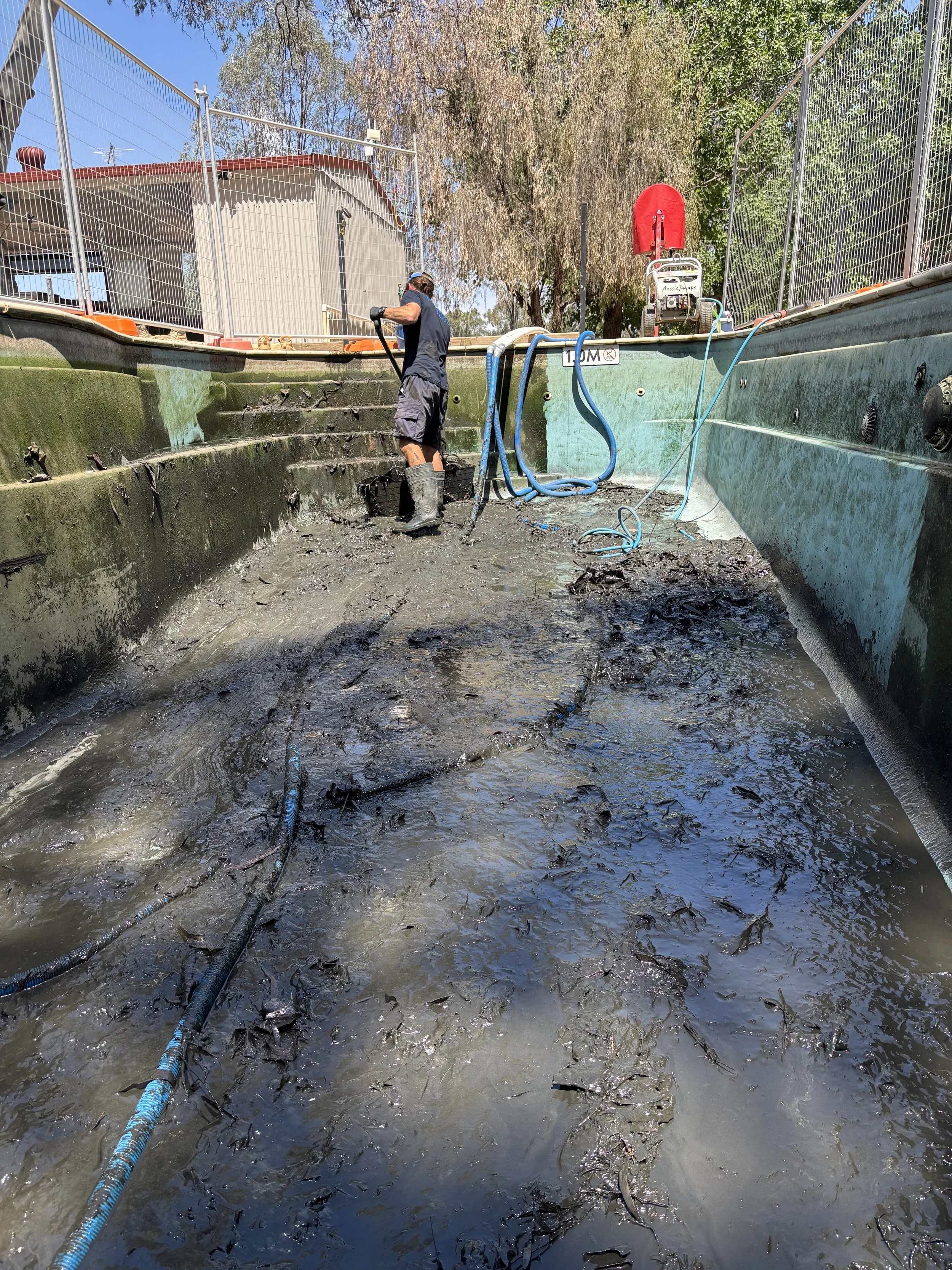 A man cleaning a muddy or algae-covered swimming pool with a high-pressure hose.