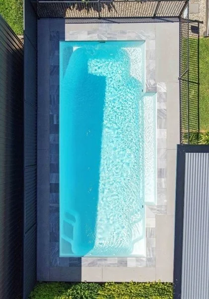 A rectangular swimming pool with clear blue water surrounded by a tiled deck and enclosed by a metal fence, viewed from above.