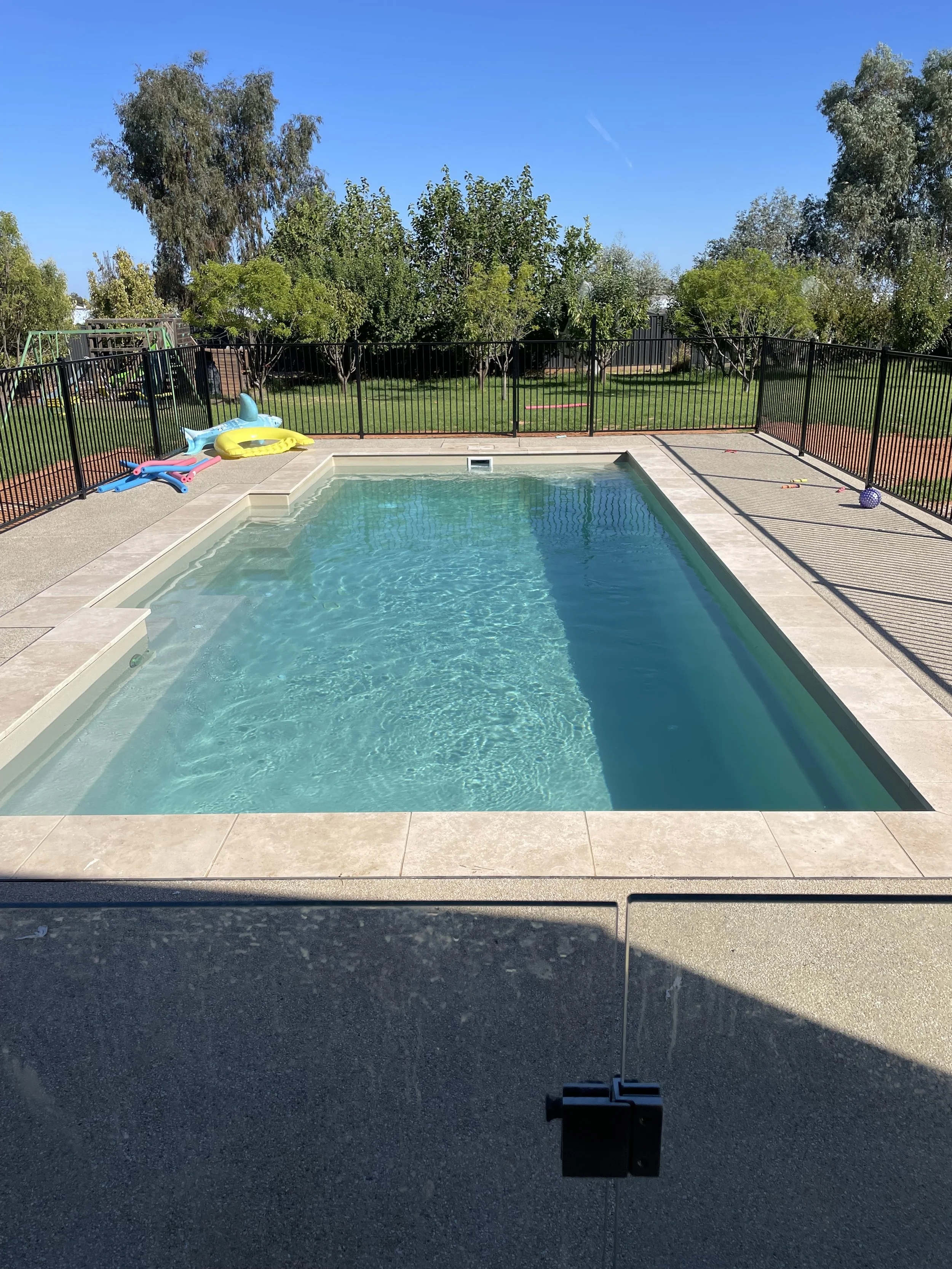 In-ground swimming pool with clear blue water, surrounded by a concrete deck, in a backyard with green grass, trees, and a black metal fence. Pool floats and toys are on the deck.