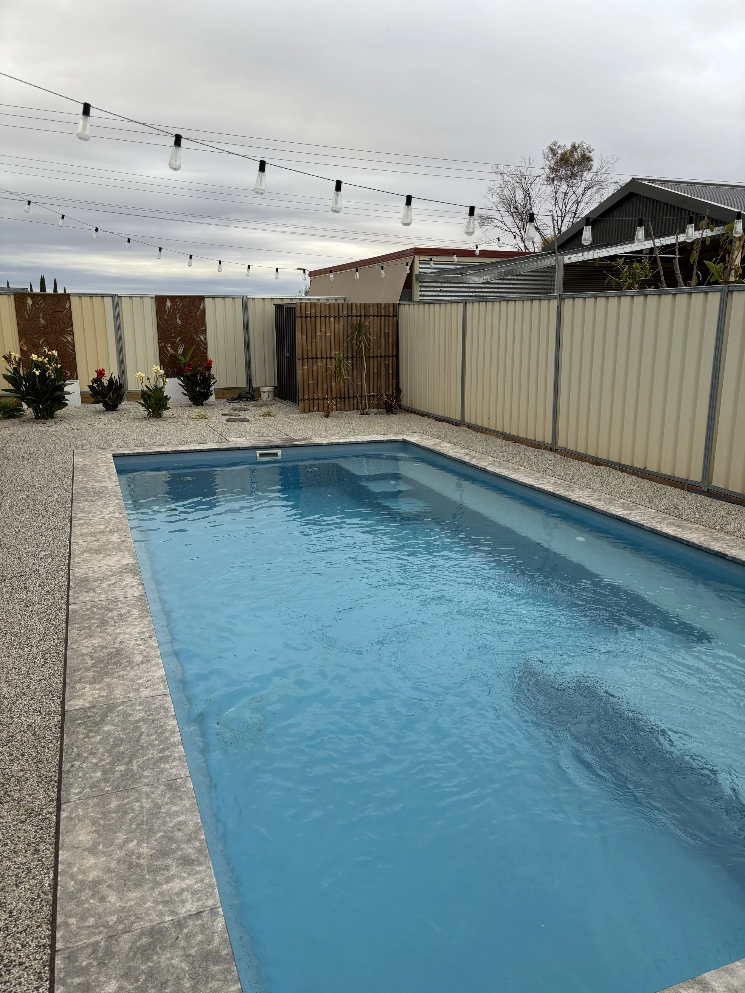 A backyard with a rectangular swimming pool, surrounded by a gravel patio, a beige fence, and a brown wooden gate. String lights are hanging overhead, and there are some plants and flowers along the fence. Overcast sky.