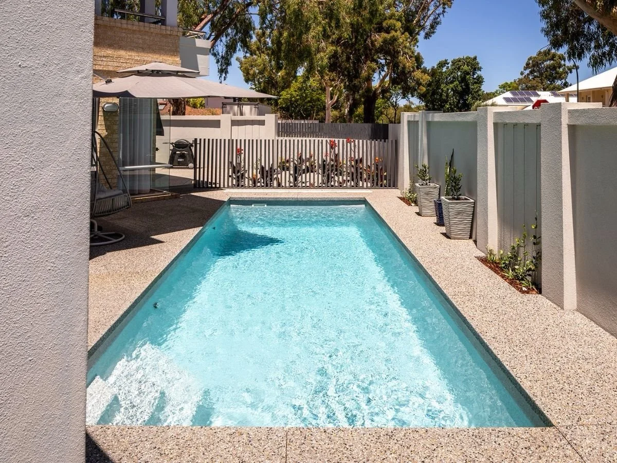 A backyard swimming pool with a textured light-colored deck, surrounded by a white fence with potted plants and trees in the background, under clear blue skies.