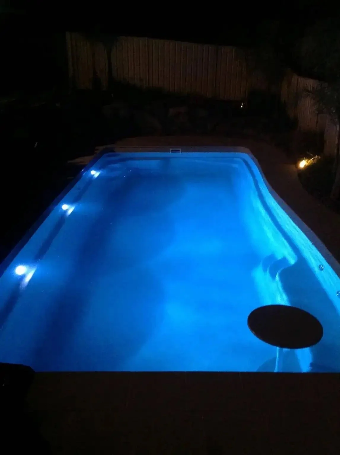 Nighttime view of a lit swimming pool in a backyard with a wooden fence and small landscape lights nearby.