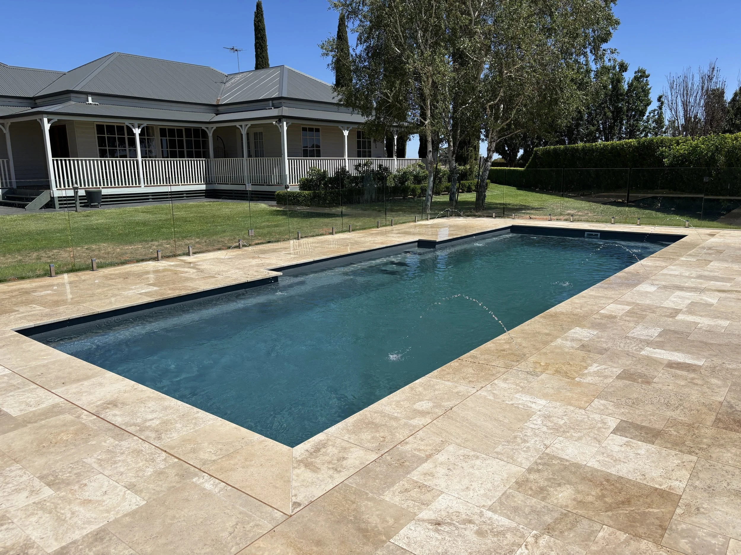 A rectangular in-ground swimming pool with beige stone tiles surrounding it, protected by a glass fence, with a house and greenery in the background under a blue sky.