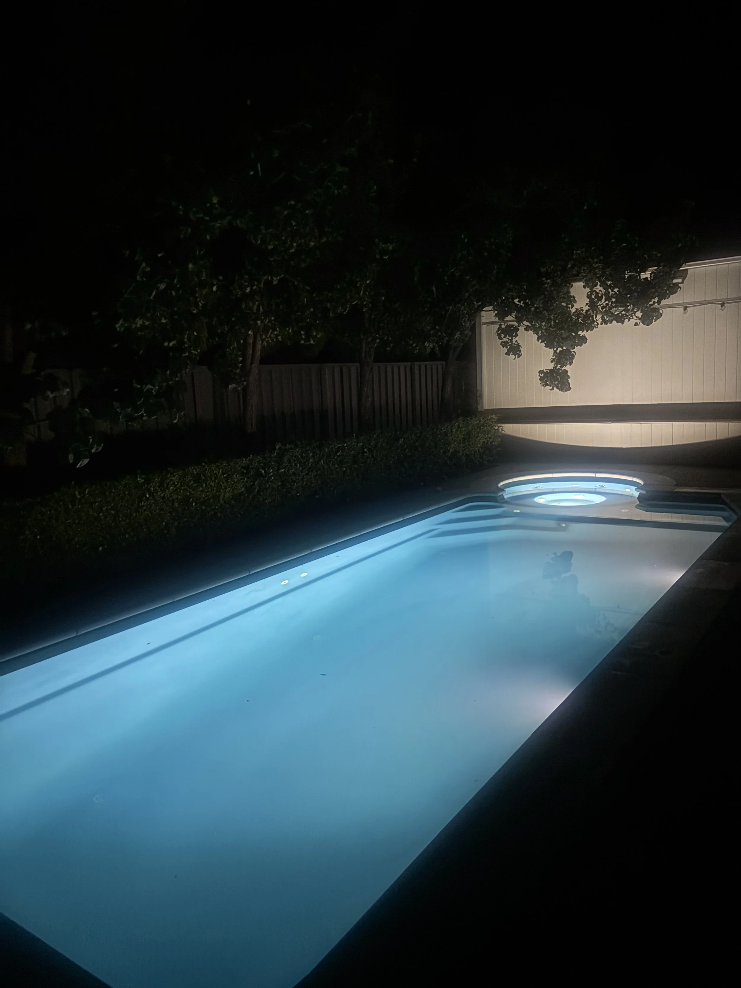 Nighttime view of a lit swimming pool with a hot tub in a backyard, surrounded by a fence and trees.