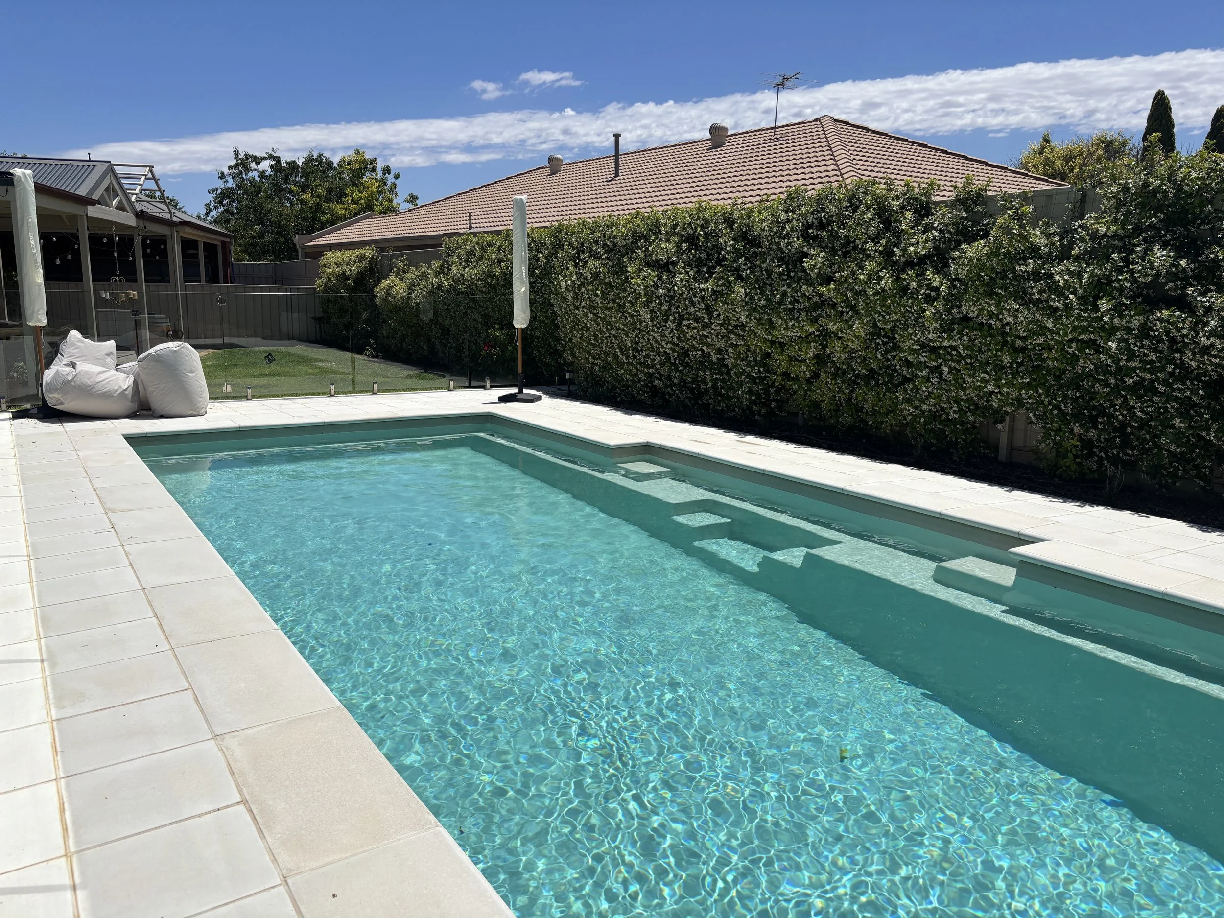 Swimming pool in backyard with steps leading into water, surrounded by white tiled deck, with two bean bags and umbrellas, fenced yard with plants and trees, blue sky with clouds.