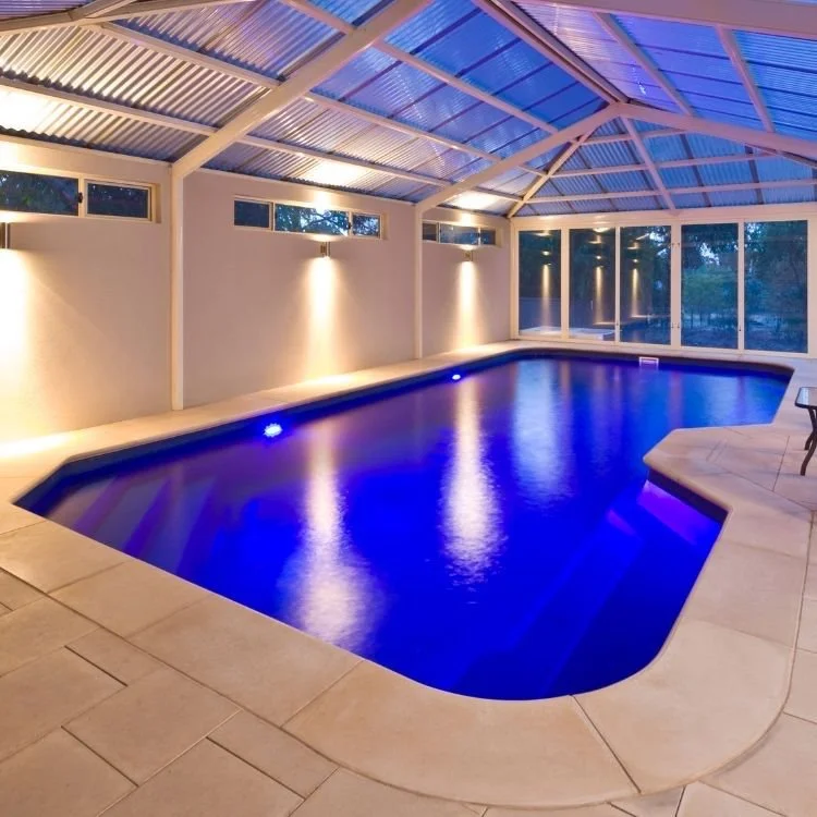 Indoor swimming pool with blue lighting, surrounded by tiled floor and enclosed with glass windows and a metal roof.