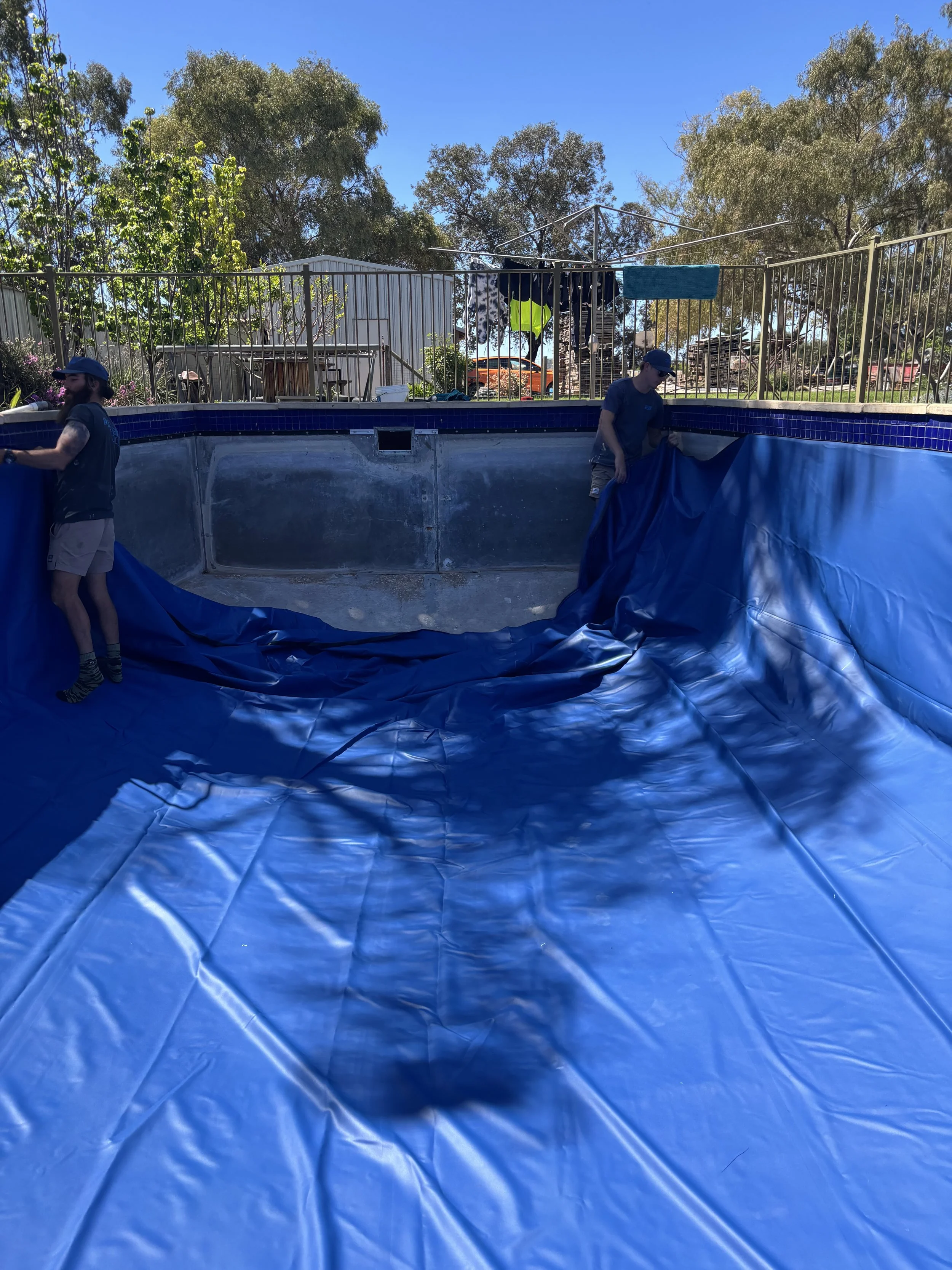 Two men installing a blue pool liner inside an empty swimming pool, with trees and a fence in the background on a sunny day.