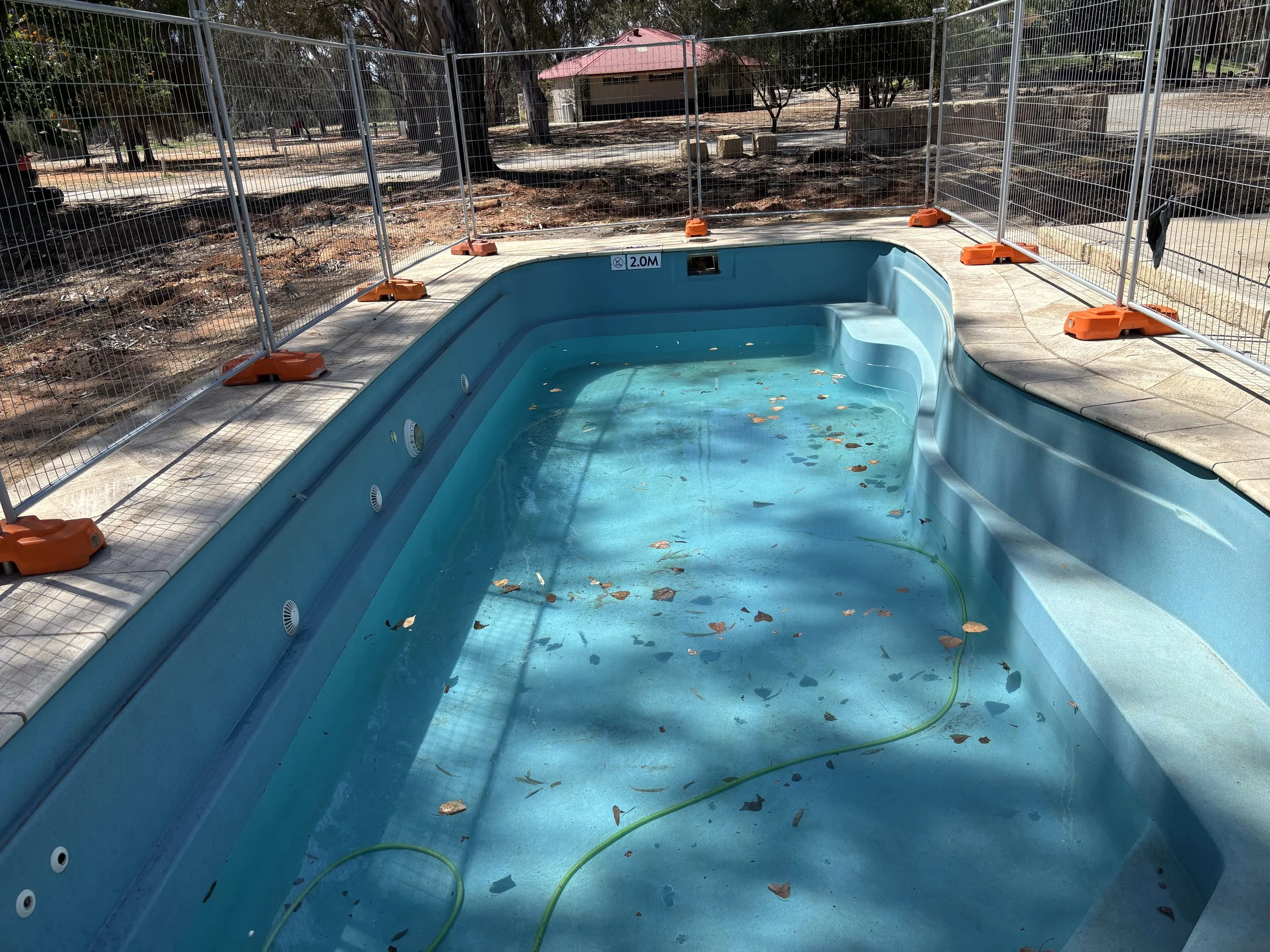 Empty swimming pool with some water, leaves, and debris inside, surrounded by a temporary metal fence.