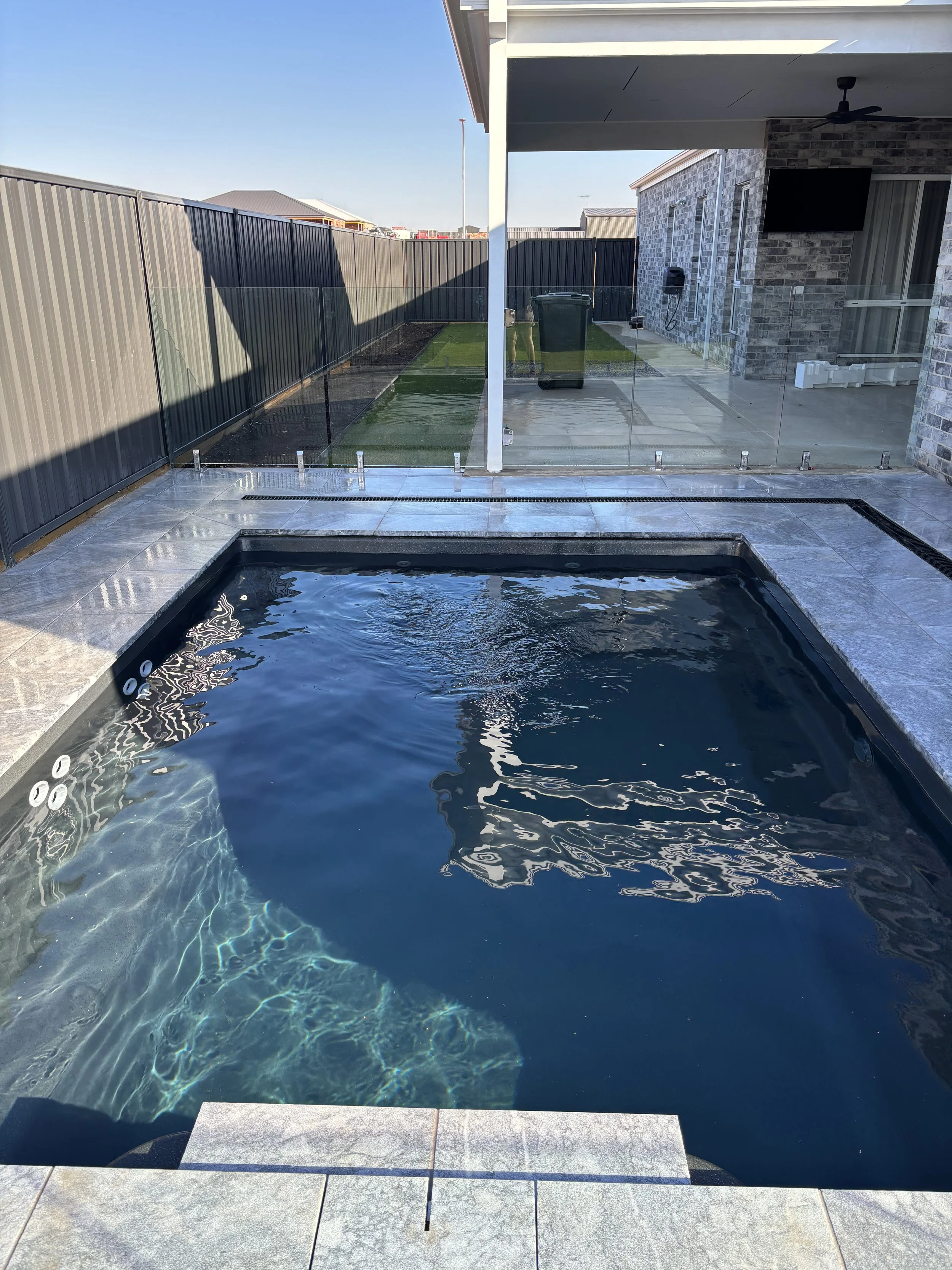 Backyard with a small in-ground swimming pool, a fenced grassy yard, and a covered patio area with a wall-mounted TV and ceiling fan.