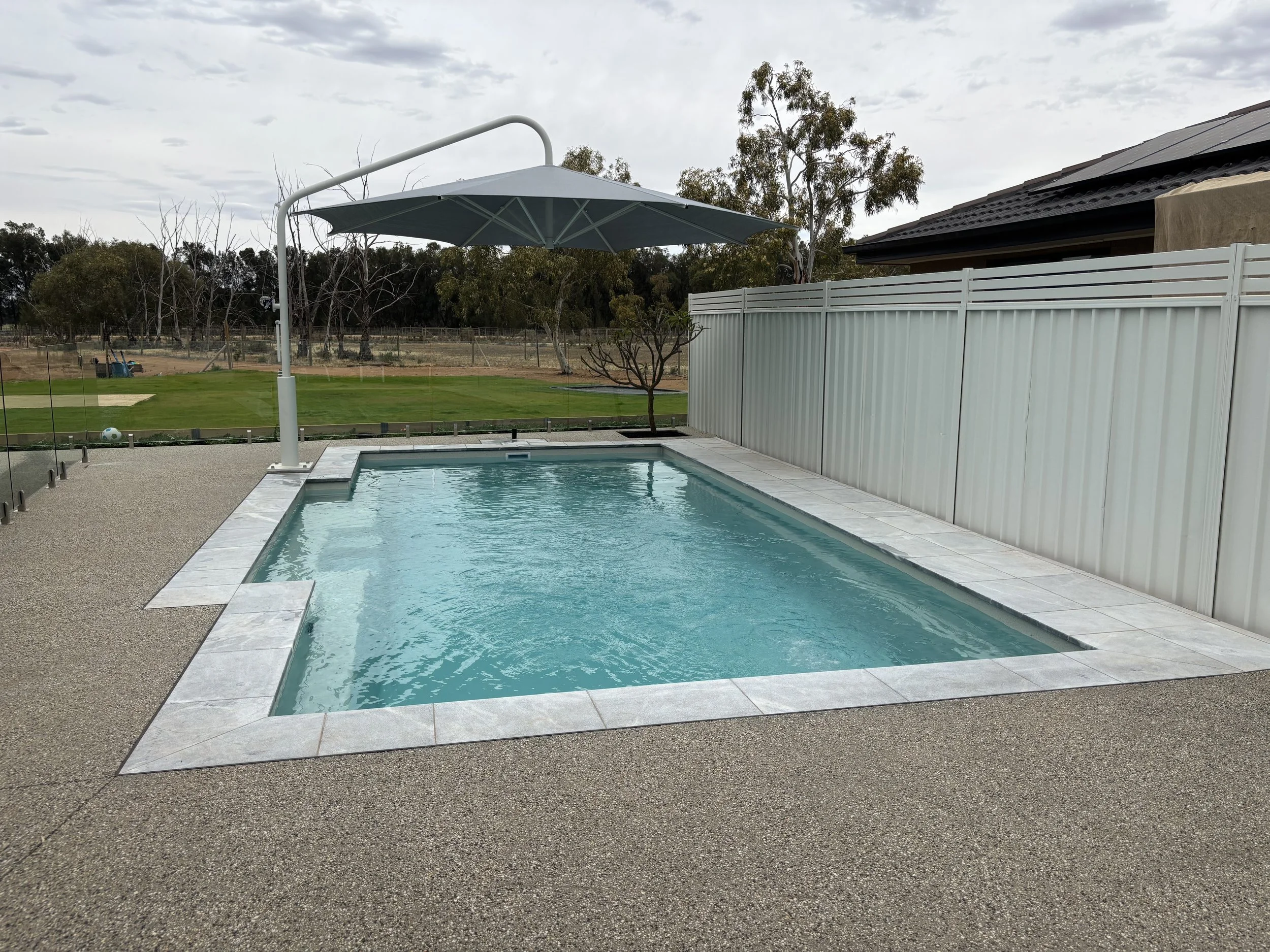 An outdoor swimming pool with a white tile border and a large gray patio umbrella over the pool, surrounded by a textured concrete deck and a white fence, with a grassy area, trees, and a partly cloudy sky in the background.