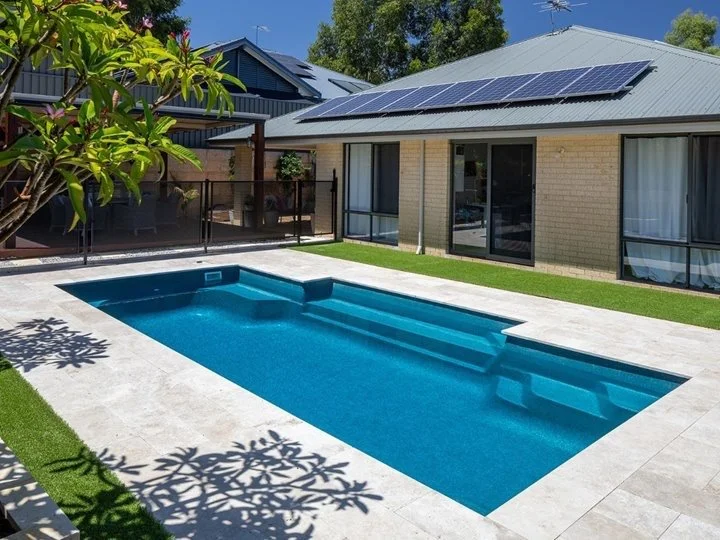 residential backyard with a blue rectangular swimming pool, surrounded by a white tile patio, a grass strip, and a house with large glass sliding doors and solar panels on the roof.
