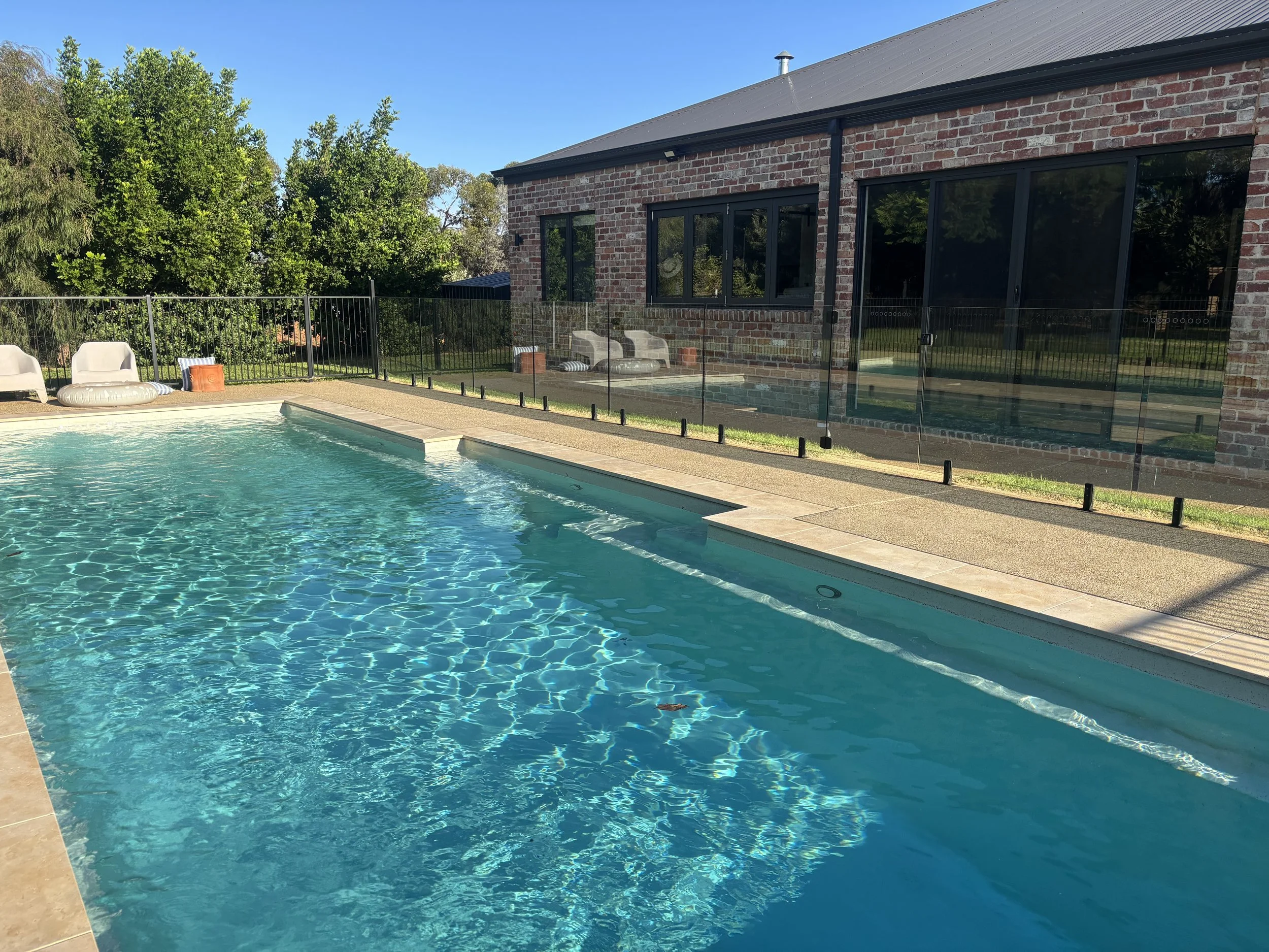 Residential backyard with a swimming pool, black fence, trees, and a brick house with large windows.