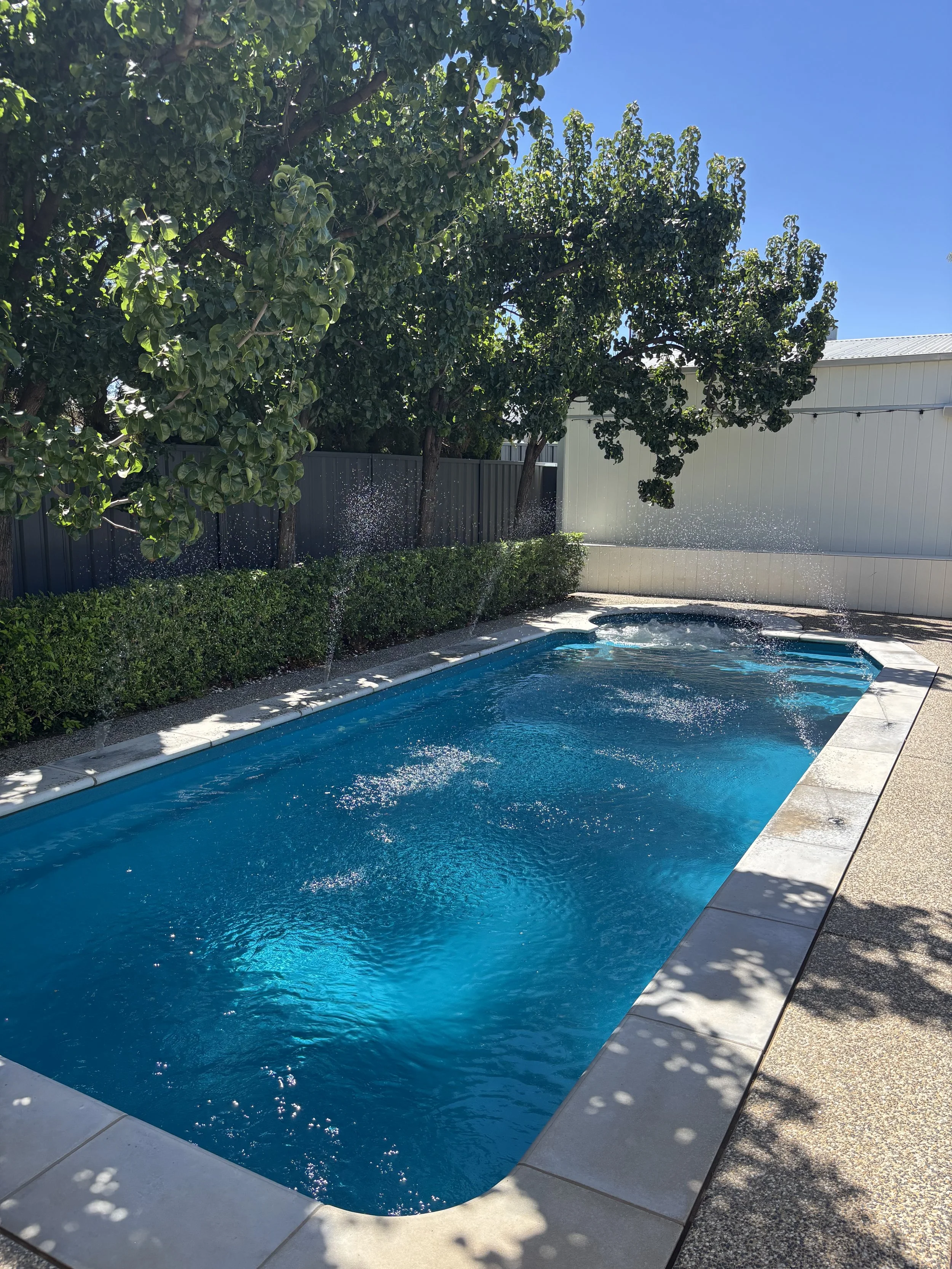 A rectangular swimming pool with clear blue water, surrounded by a paved deck, nearby trees providing partial shade, and a white building and fence in the background under a blue sky.