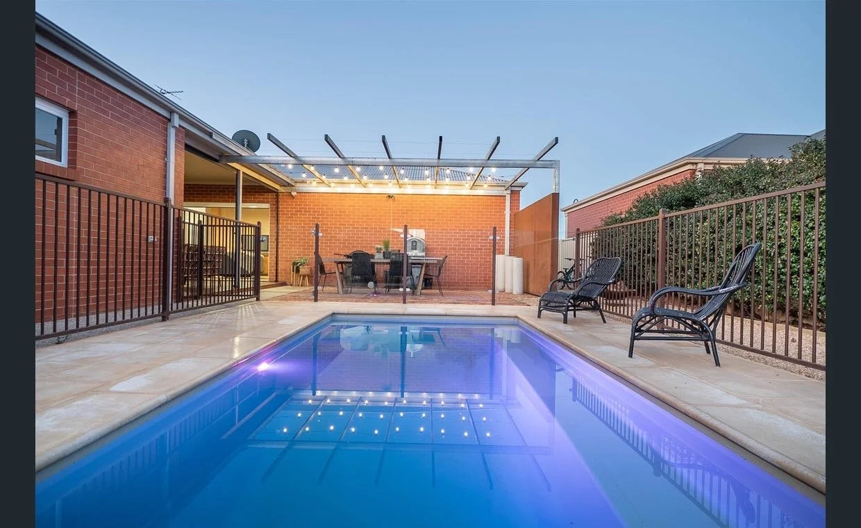 A backyard with a small swimming pool, surrounded by a metal fence. There are two black patio chairs on the patio near the pool. In the background, there is a brick house with outdoor dining furniture and a grill under a pergola with string lights.