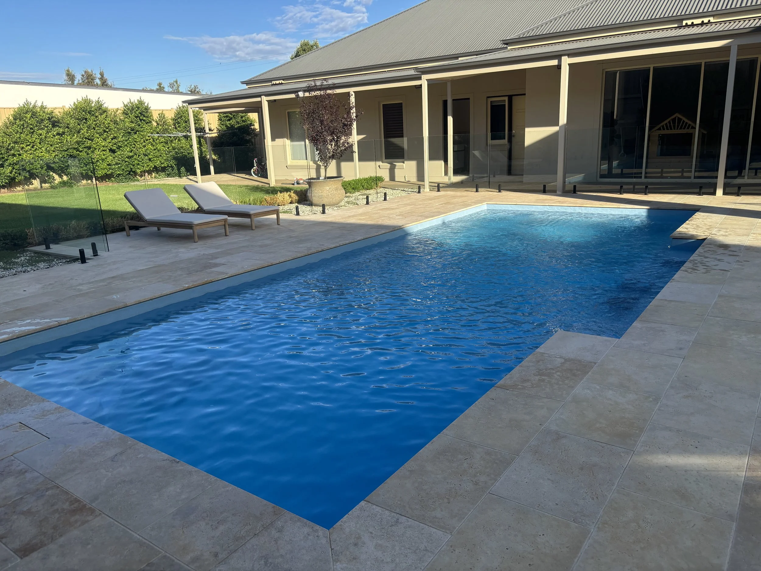 A backyard with a rectangular swimming pool, two lounge chairs, and a house with a covered patio. There is a potted tree and a glass fence around part of the pool area. Green trees and a blue sky are in the background.