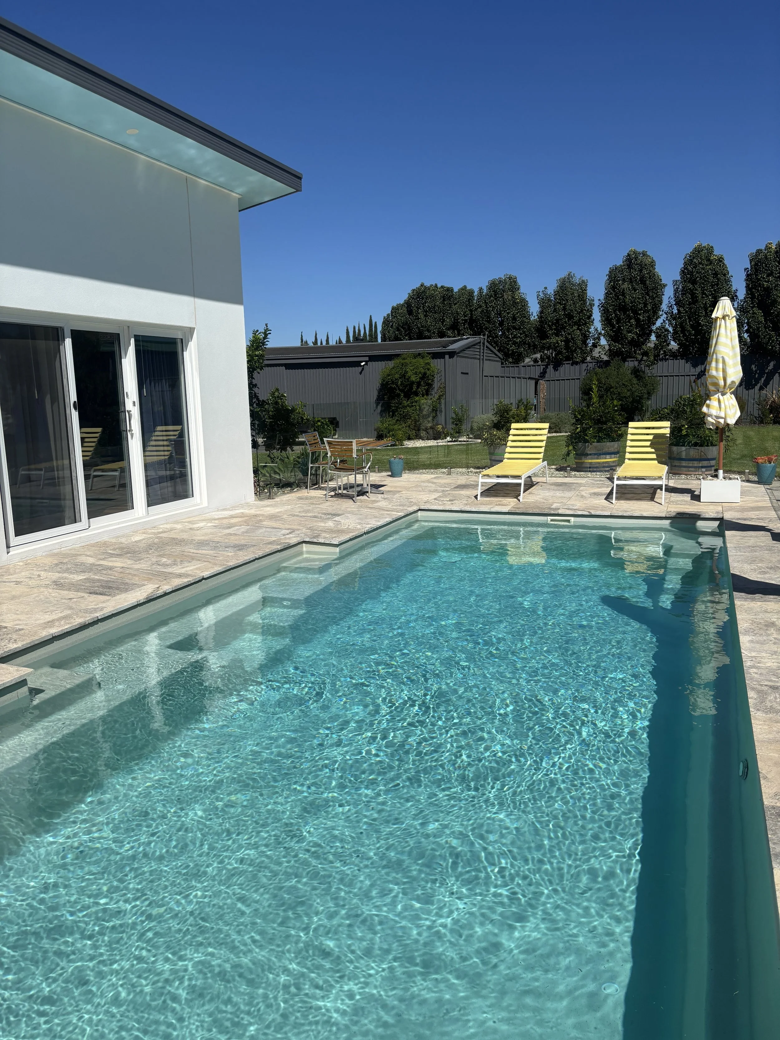 A backyard swimming pool area with yellow lounge chairs, a striped patio umbrella, and a modern house with sliding glass doors under a clear blue sky.
