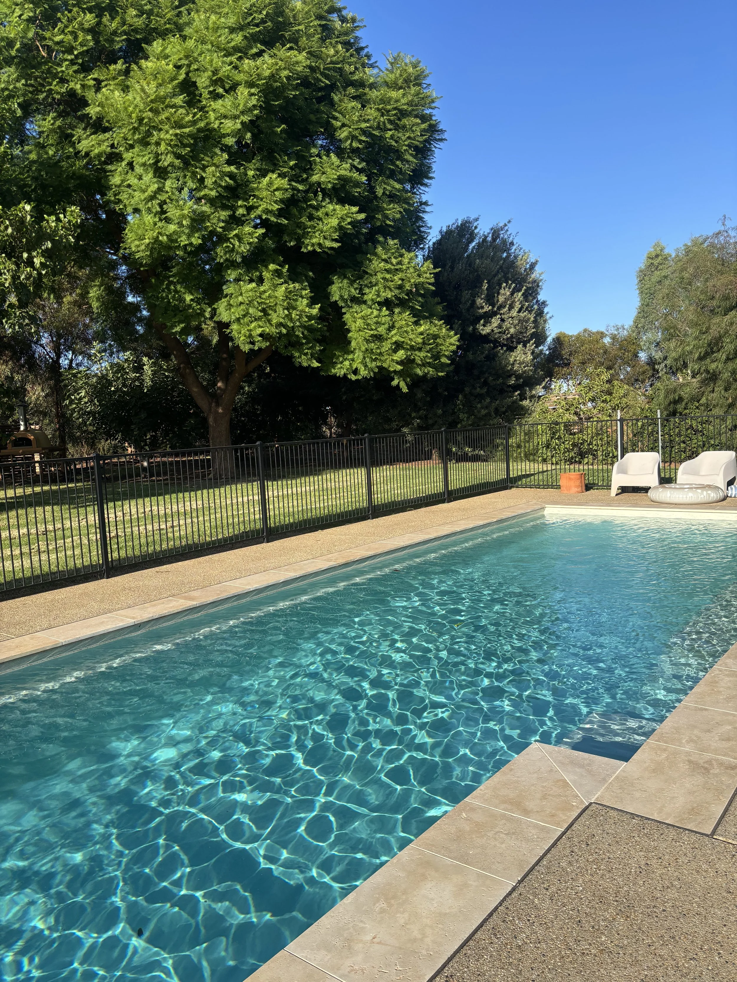 An outdoor swimming pool with clear blue water, surrounded by a concrete deck and black metal fence, with green trees and a blue sky in the background.