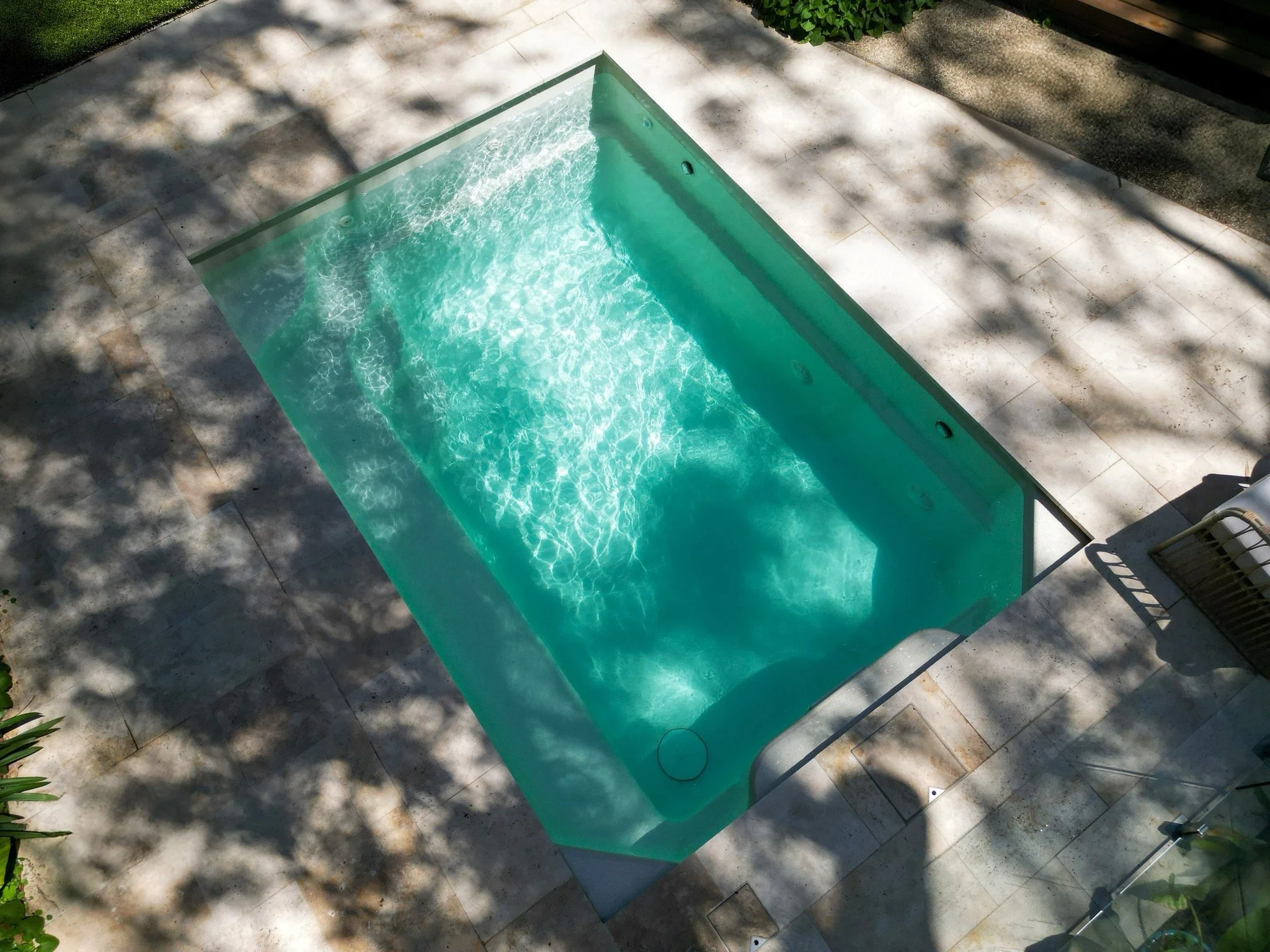 A rectangular outdoor hot tub with bubbling water, surrounded by a tiled patio with shadows from nearby trees.