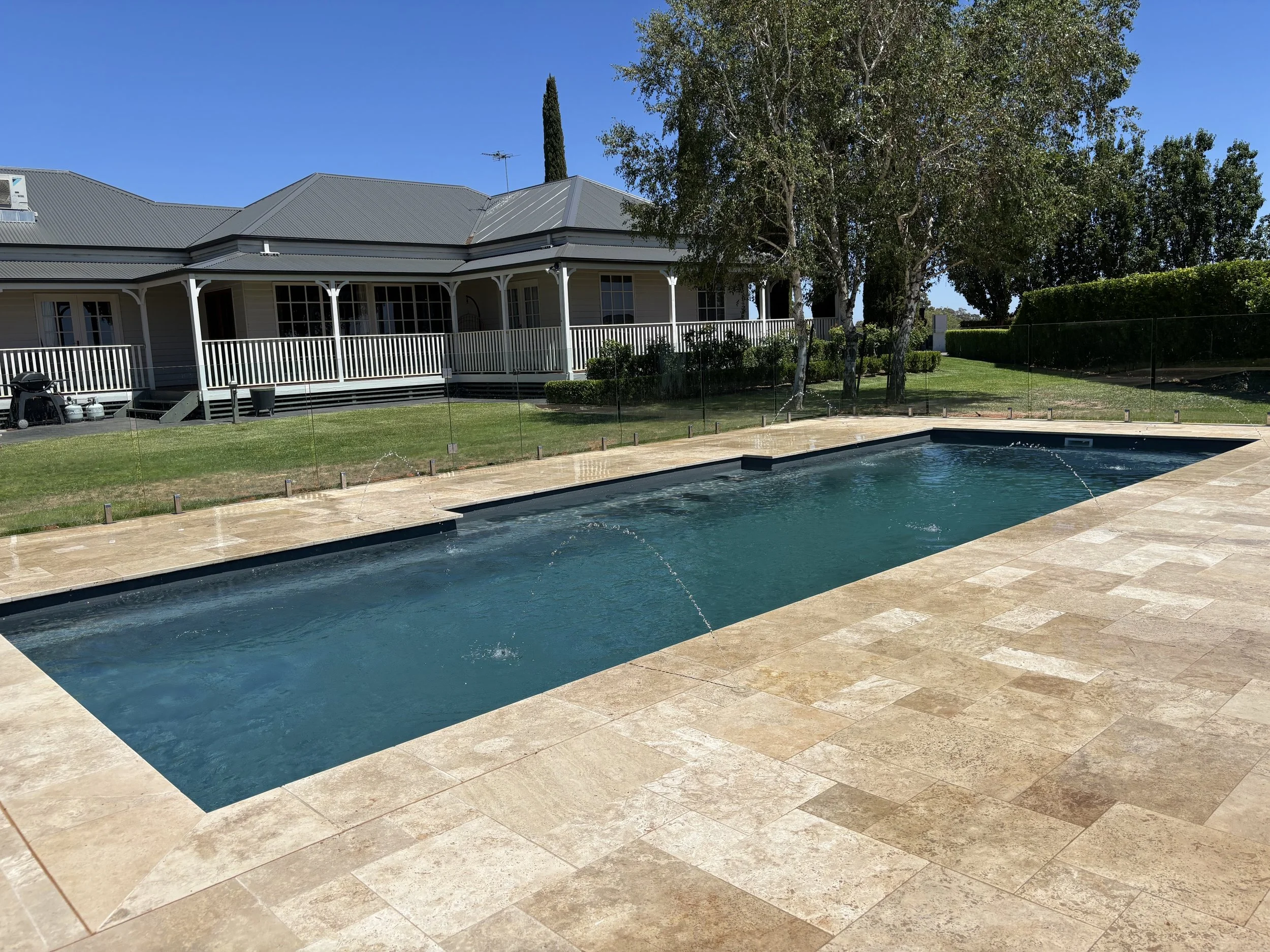 Residential backyard with a rectangular swimming pool, beige stone patio, green lawn, trees, and a house with a gray metal roof and porch in the background under a clear blue sky.