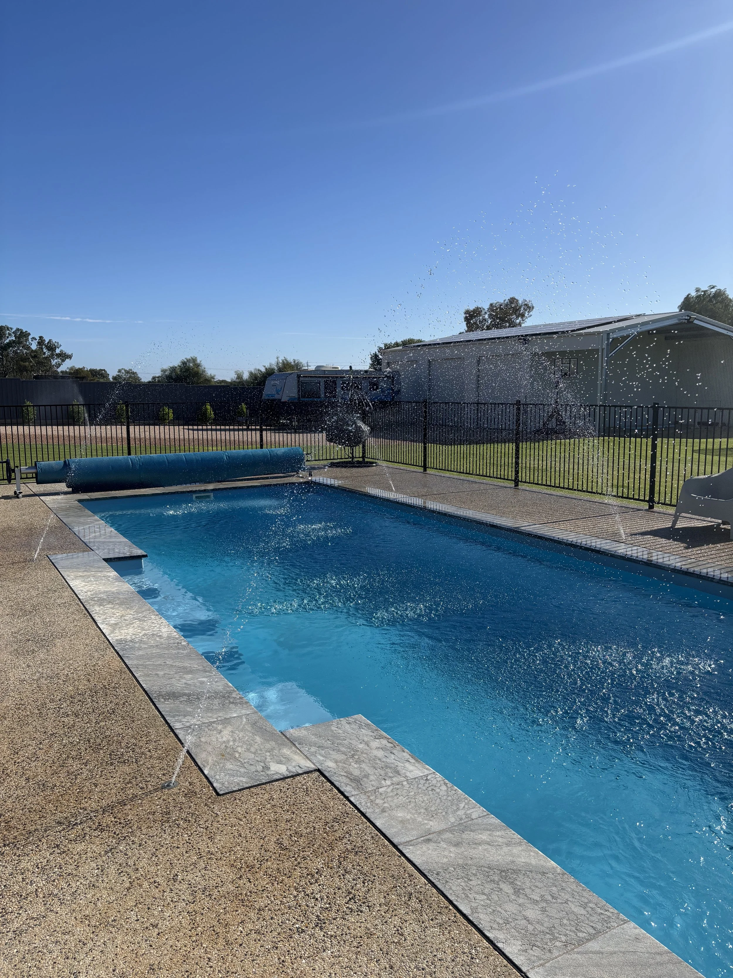 A backyard swimming pool with a safety fence, blue water, and a pool cover rolled up on the edge. In the background, there is a grassy area with trees, a white shed or garage, and a recreational vehicle (RV). The sky is clear and sunny.