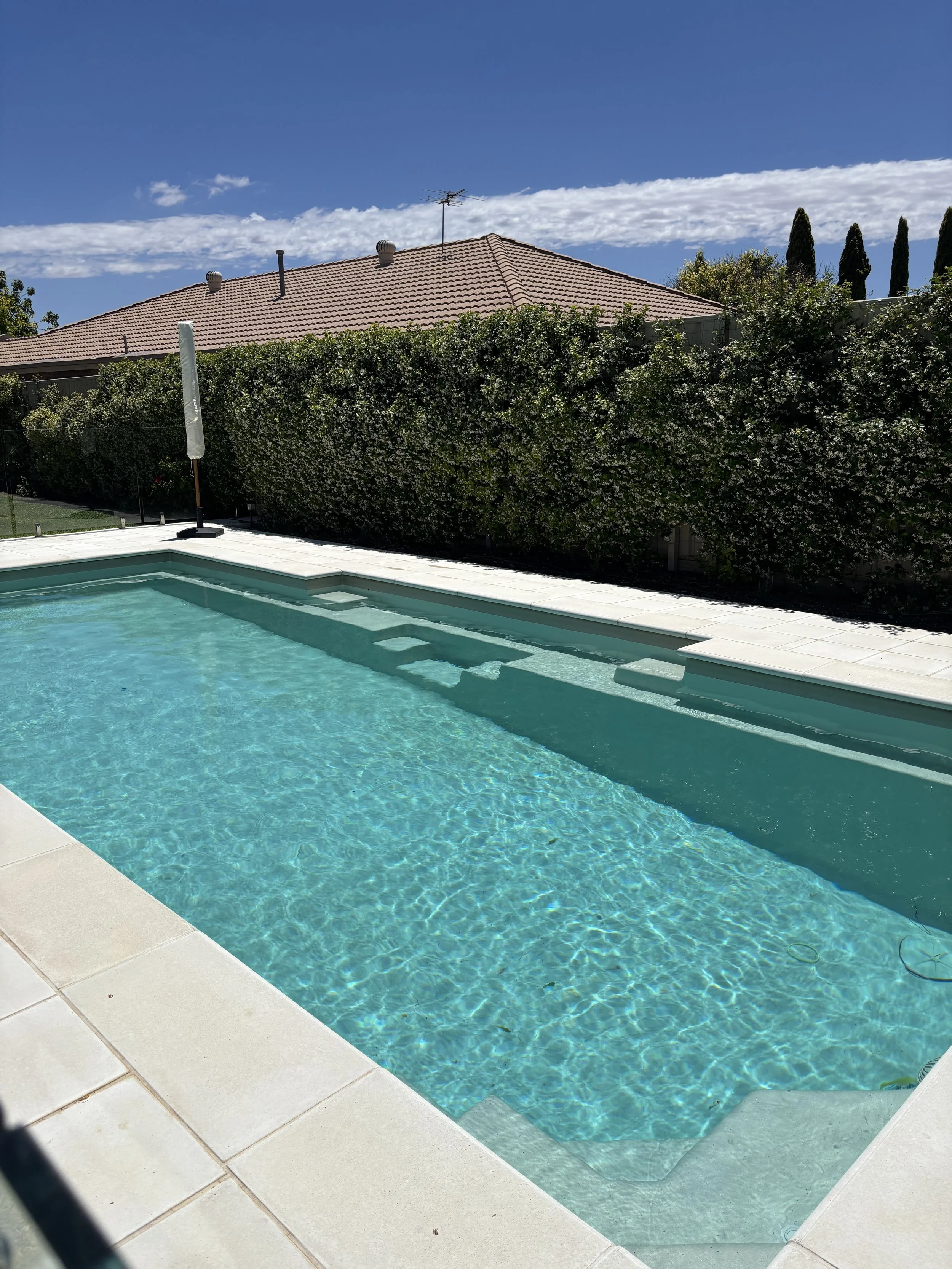 Backyard swimming pool with clean water, surrounded by white tile deck, with a tall hedge and house with a tiled roof in the background, under a partly cloudy sky.