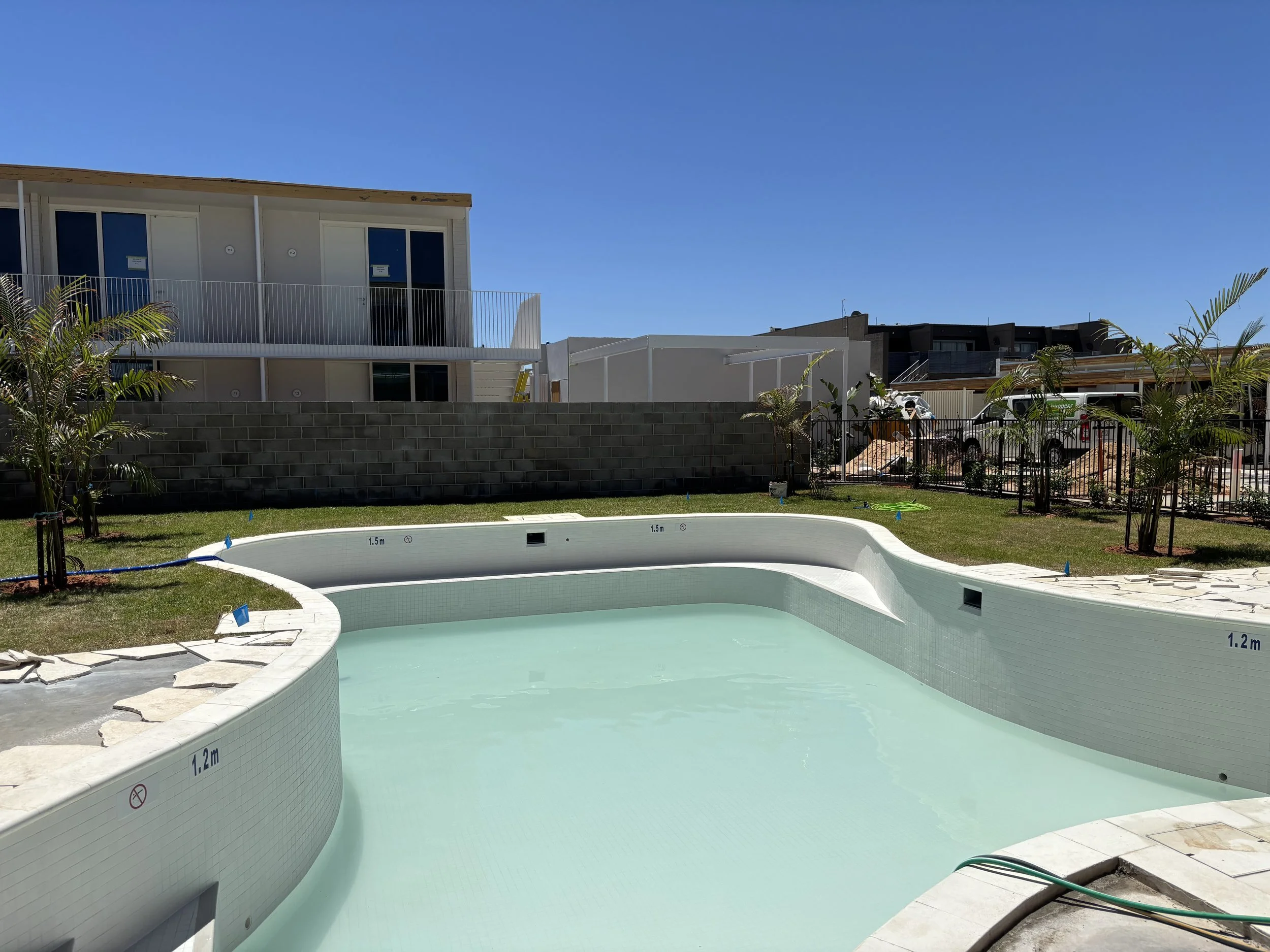 Empty swimming pool under construction in a backyard with a grassy area, small trees, and new residential buildings in the background under a clear blue sky.