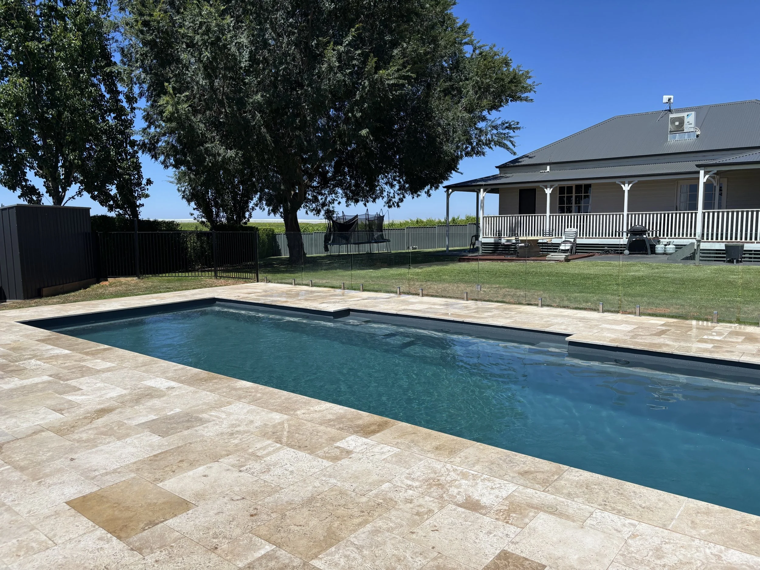 Backyard with in-ground swimming pool surrounded by a stone patio, enclosed by a glass fence. A large tree and a house with a porch are visible in the background under a clear blue sky.