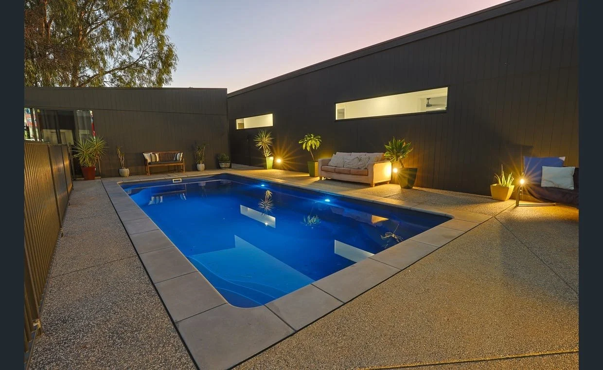 Nighttime view of a backyard with a rectangular swimming pool illuminated by underwater lights, surrounded by potted plants and outdoor furniture, with a black modern building wall and string lights.