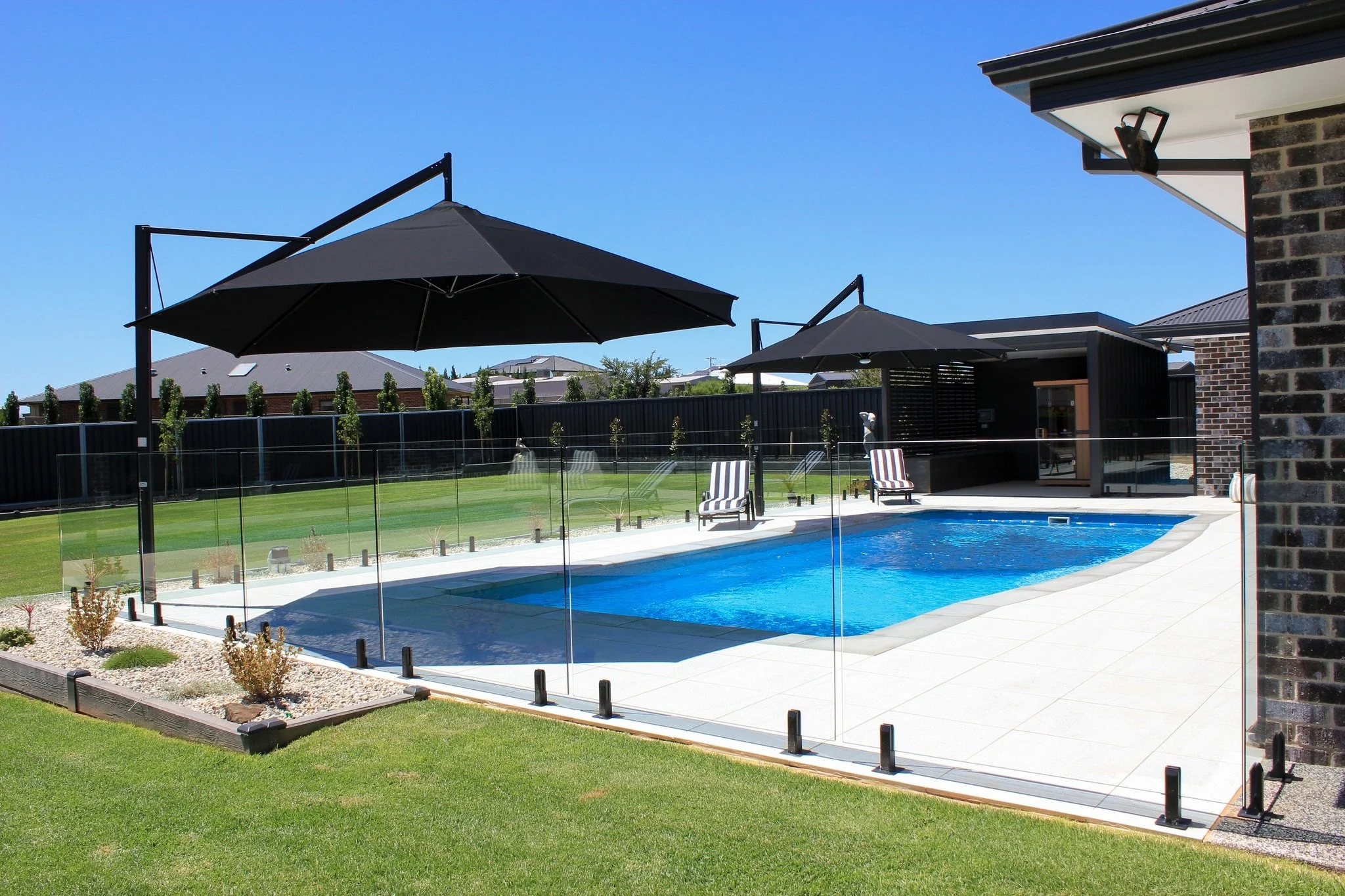 A backyard with a swimming pool enclosed by glass panels, surrounded by patio chairs, large umbrellas, and a well-maintained lawn with a black fence and trees in the background.