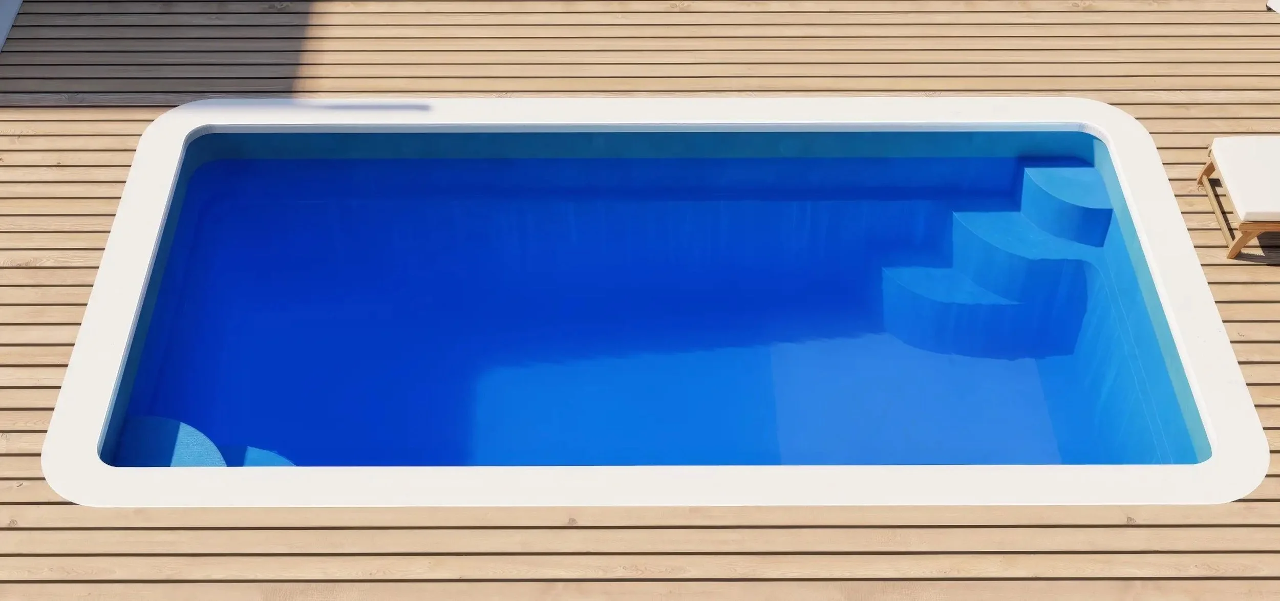 An empty swimming pool with blue water, surrounded by wooden decking and a lounge chair nearby.